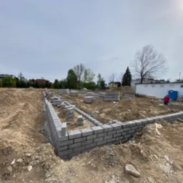 Wykop pod fundamenty z częściowo wymurowanymi ścianami z szarego bloczka betonowego; widoczny teren budowy, drzewa w tle i budynek gospodarczy.