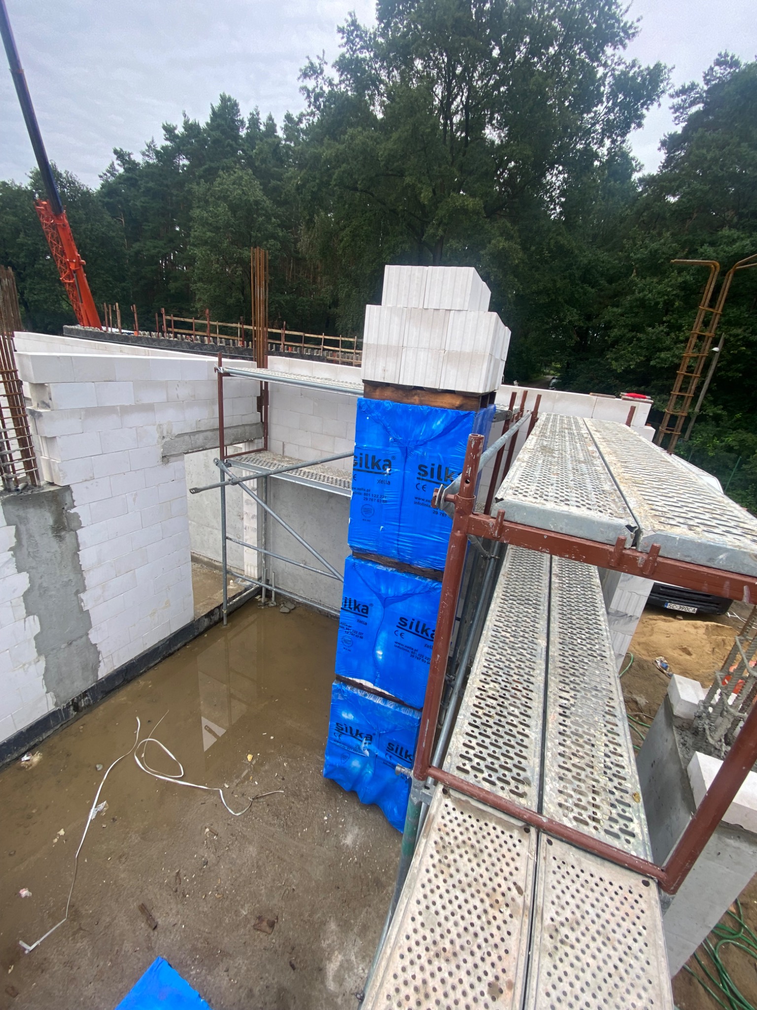 Budowa domu z białych bloczków Silka, widoczne ściany w stanie surowym, rusztowanie, paleta bloczków w niebieskiej folii, teren budowy po deszczu, żuraw budowlany w tle.