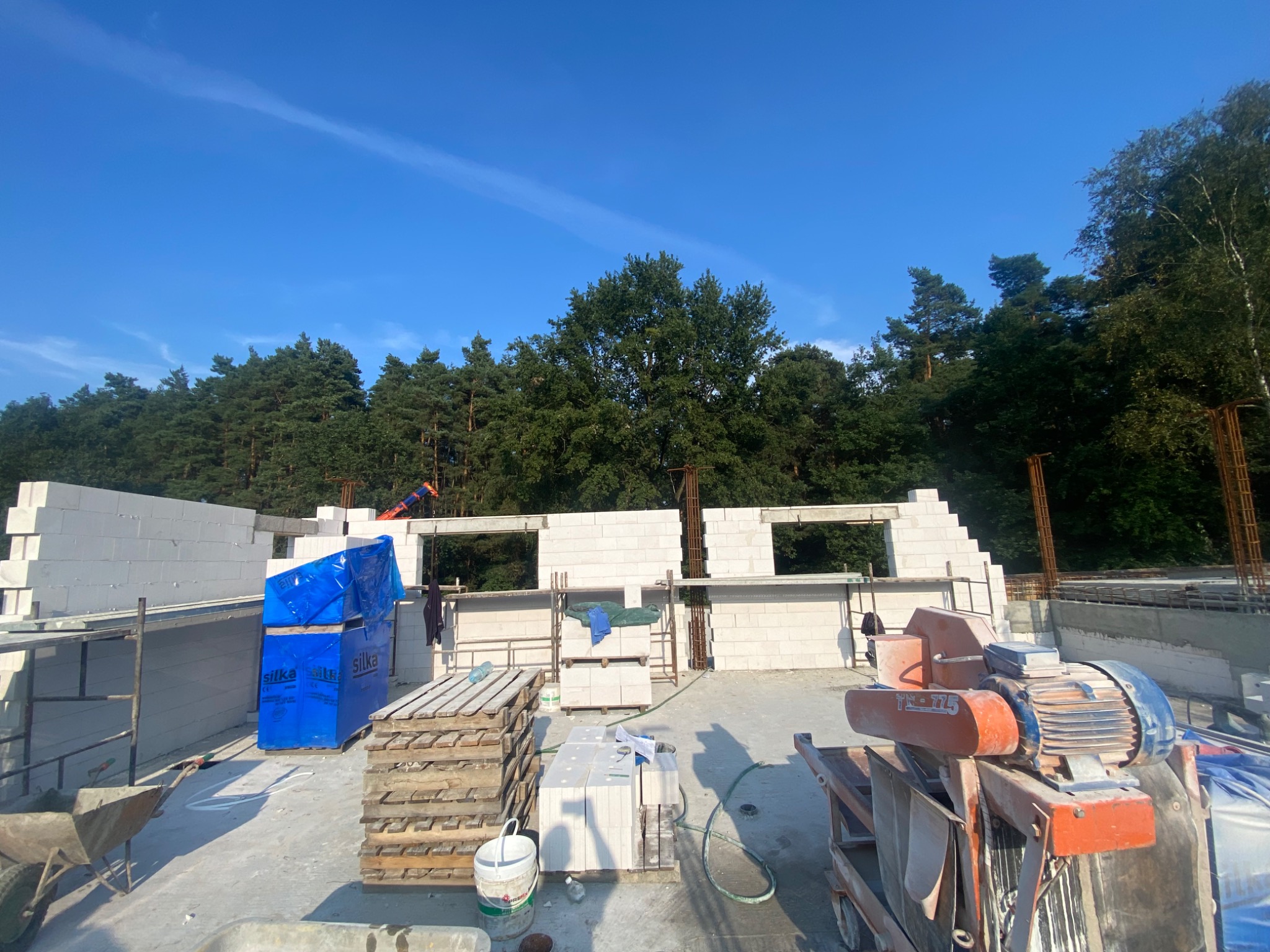 Wznoszenie ścian domu z bloczków Silka, widoczna paleta zafoliowanych bloczków, mieszadło do zaprawy, zbrojenie i drzewa w tle pod błękitnym niebem.