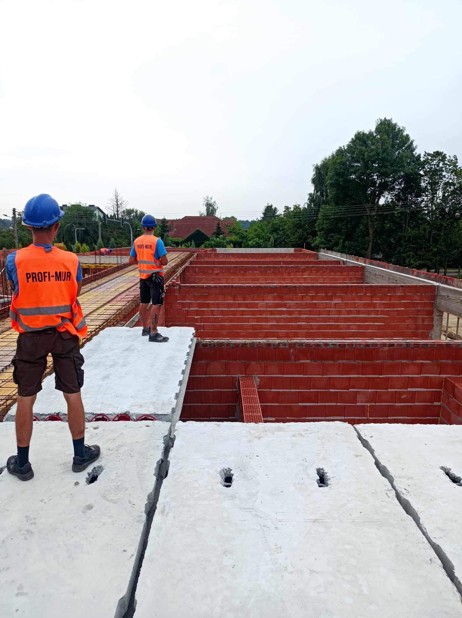 Dwóch pracowników w kaskach i pomarańczowych kamizelkach firmy PROFI-MUR nadzoruje budowę ścian z czerwonej cegły, stojąc na prefabrykowanych elementach stropowych z otworami.