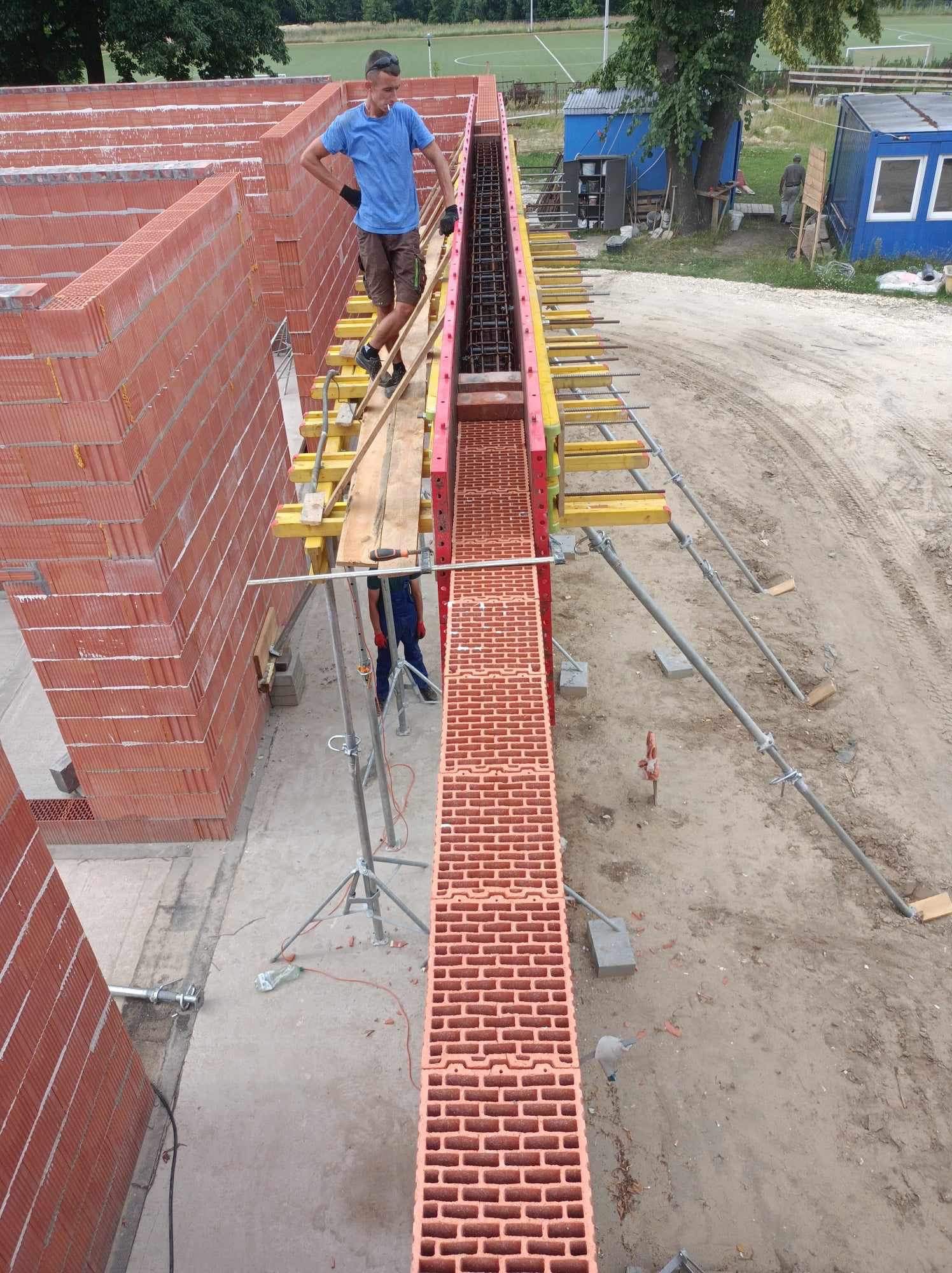 Wysoki kąt widzenia na budowę z czerwonej cegły z pracownikiem stojącym na rusztowaniu, widoczne zbrojenie i teren budowy z barakami w tle.