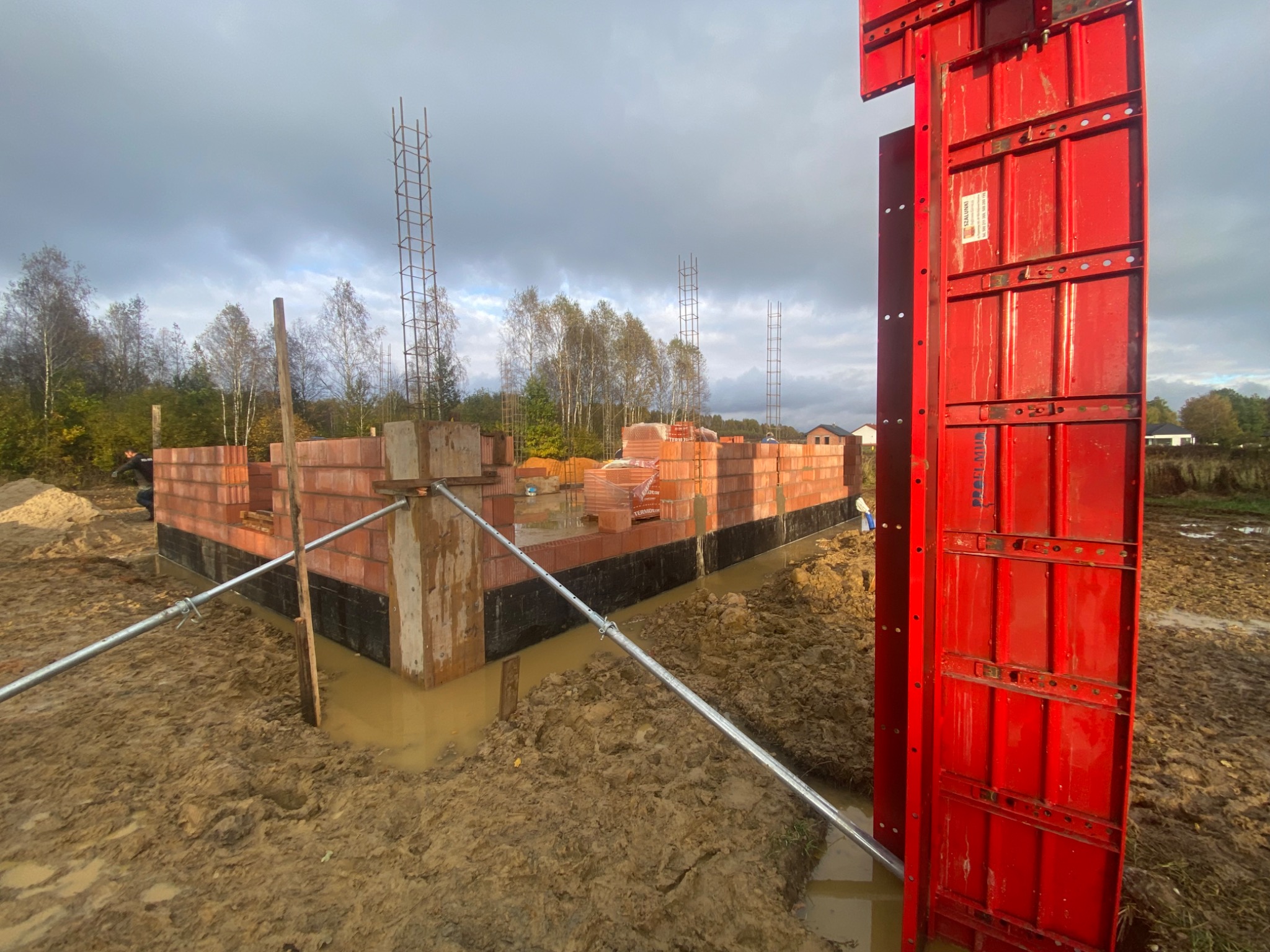 Wznoszenie ścian domu z czerwonej cegły, widoczne szalunki w kolorze czerwonym, stalowe pręty zbrojeniowe wystające pionowo, teren wokół w stanie surowym, błotnisty.