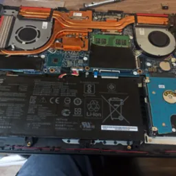 Wnętrze otwartego laptopa ASUS z widoczną baterią, układem chłodzenia z miedzianymi rurkami cieplnymi, wentylatorami, dyskiem HDD i pamięcią RAM. Widoczne ślady demontażu i drobne zabrudzenia.