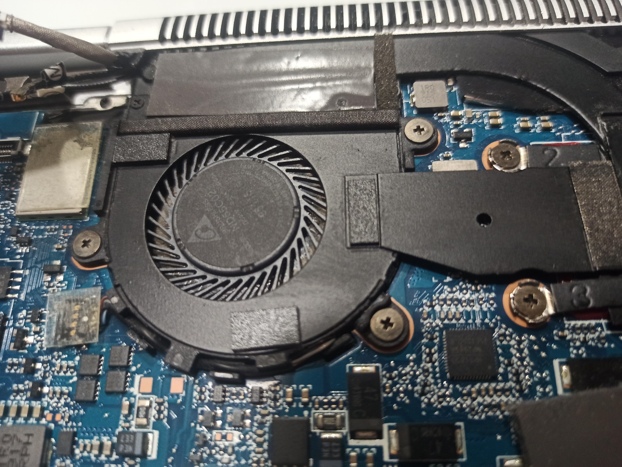 Wnętrze laptopa z widocznym wentylatorem chłodzącym, płyta główna z elementami elektronicznymi, śrubki mocujące elementy, widoczne oznaczenia na wentylatorze