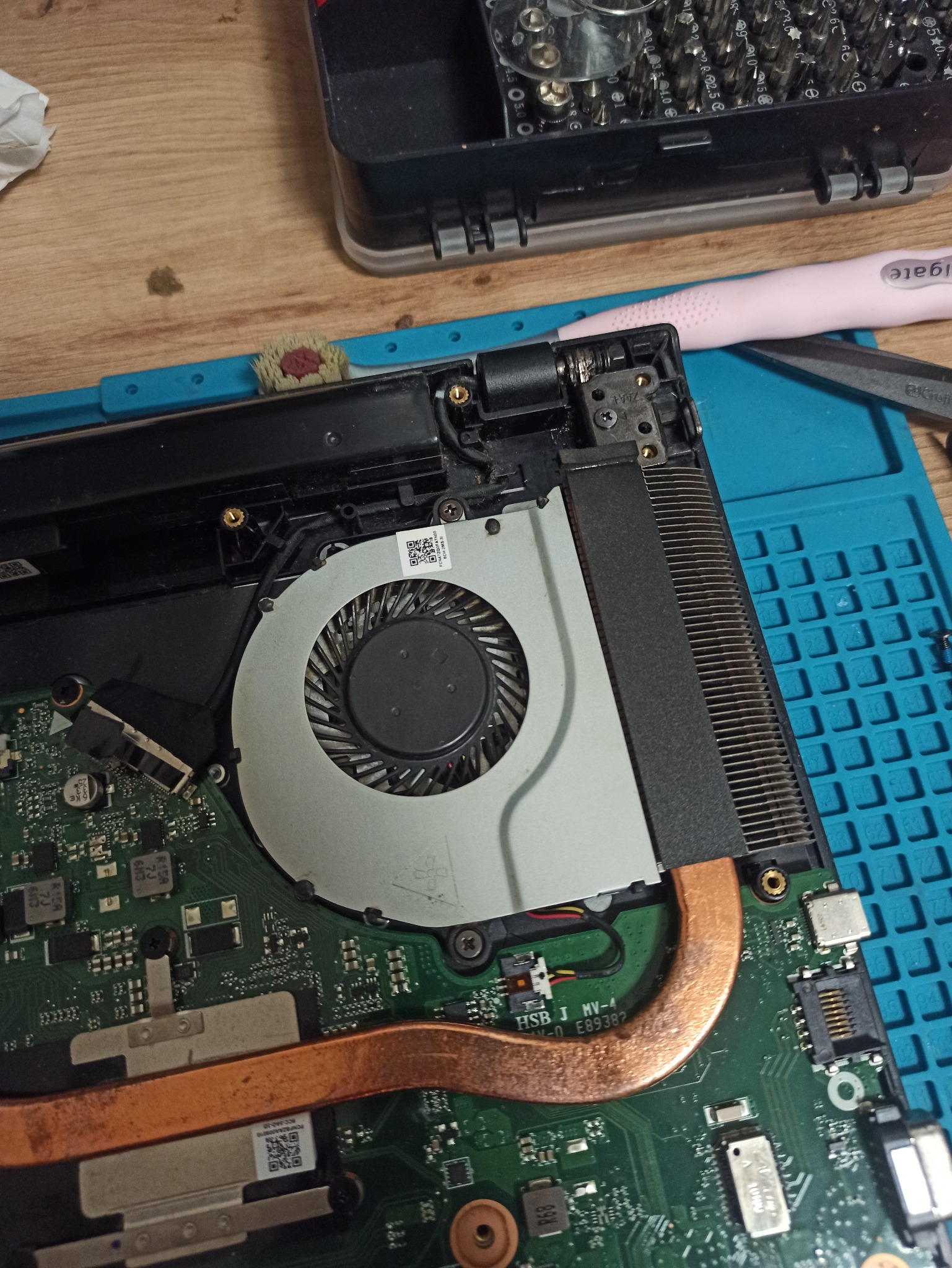 Wnętrze laptopa z widocznym układem chłodzenia, wentylatorem i elementami płyty głównej na niebieskiej macie serwisowej, obok narzędzia i pojemnik z bitami.