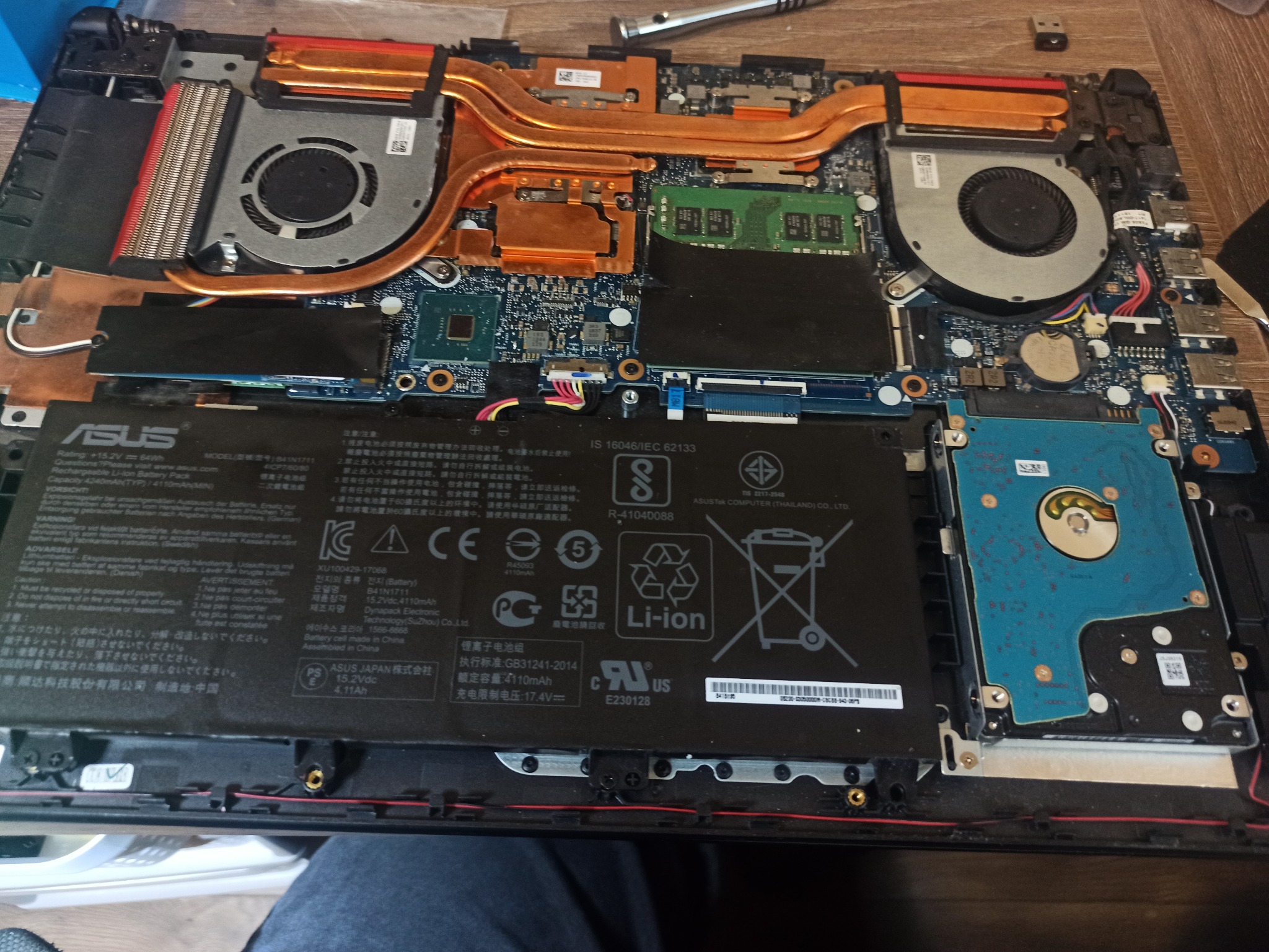 Wnętrze otwartego laptopa ASUS z widoczną baterią, układem chłodzenia z miedzianymi rurkami cieplnymi, wentylatorami, dyskiem HDD i pamięcią RAM. Widoczne ślady demontażu i drobne zabrudzenia.