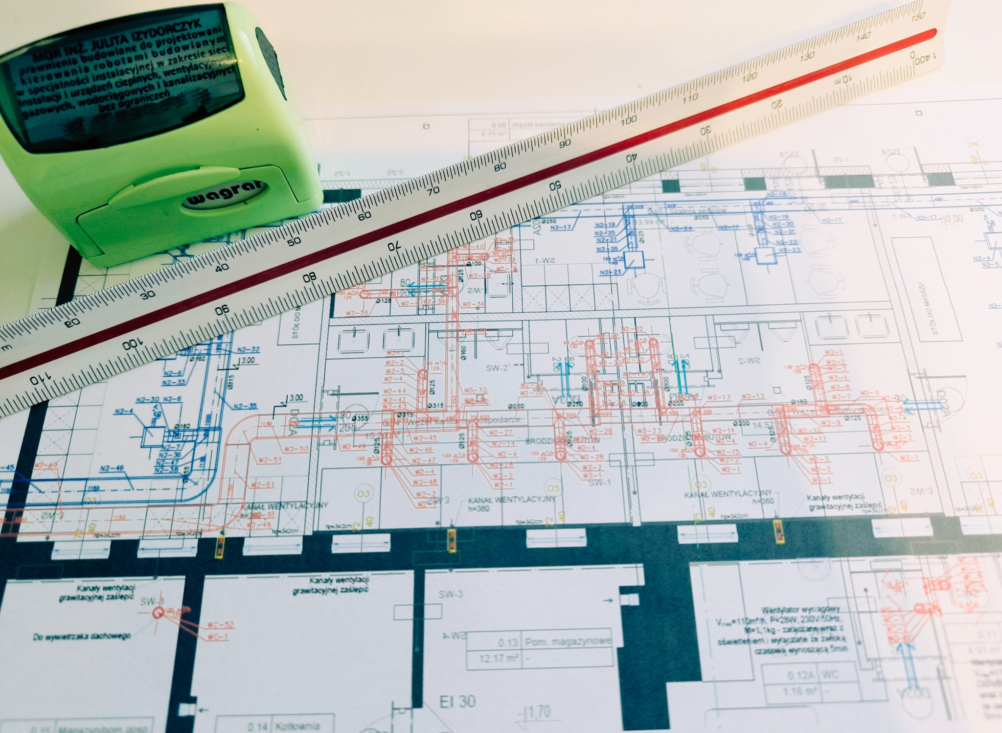 Plan instalacji sanitarnych z naniesionymi oznaczeniami, przykryty częściowo przez zielony stempel z danymi projektanta i białą linijkę z podziałką centymetrową.