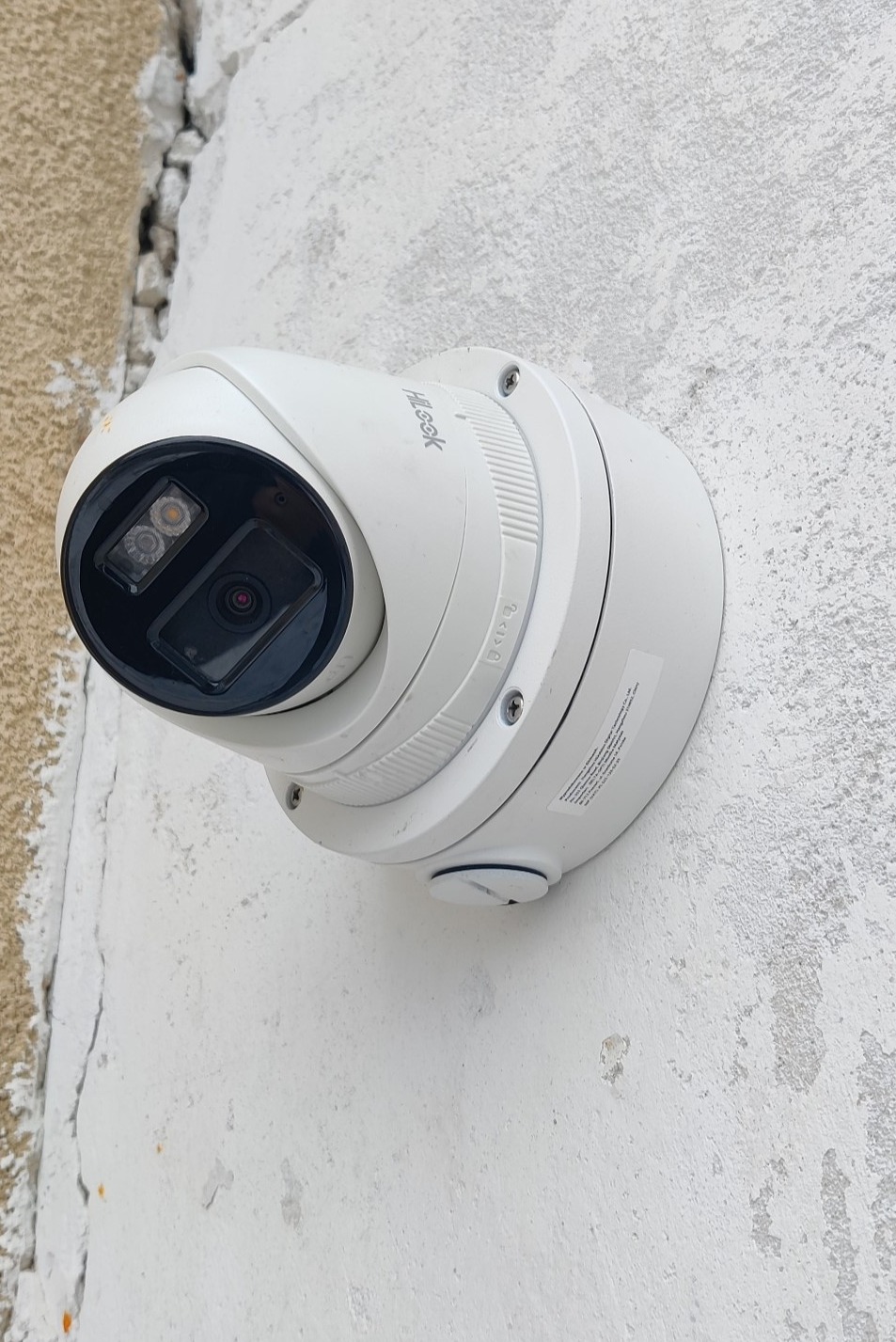 Kamera monitoringu marki Hilook zamontowana na białej, lekko uszkodzonej ścianie w Białymstoku. Widoczne śruby mocujące i fragment etykiety informacyjnej na obudowie.