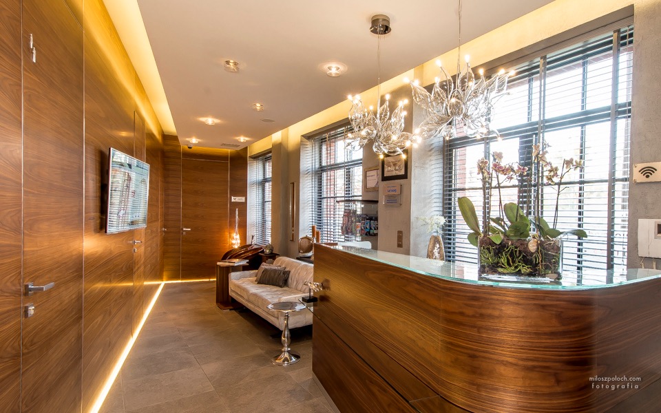 Nowoczesne wnętrze recepcji z drewnianą ladą, kryształowymi żyrandolami i sofą w tle, doświetlone ledowym oświetleniem wzdłuż ściany.