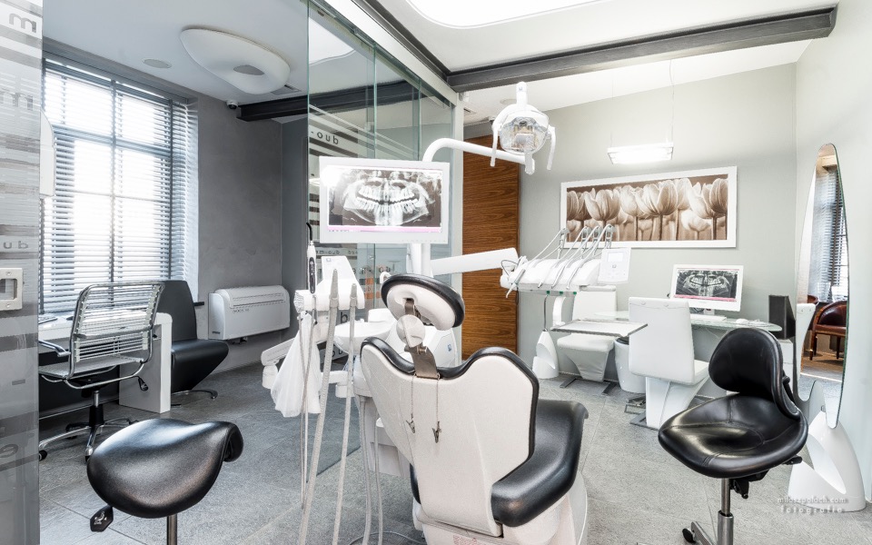 Nowoczesny gabinet stomatologiczny z panoramicznym zdjęciem zębów na monitorze, wyposażony w biały fotel dentystyczny i lampę, z szarymi ścianami i subtelnym oświetleniem