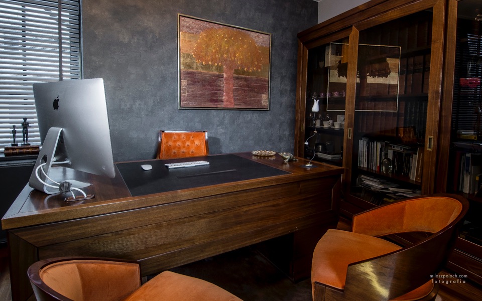 Eleganckie biuro z drewnianym biurkiem, komputerem Apple, pomarańczowym krzesłem i obrazem drzewa na ścianie, uzupełnione regałem z książkami.