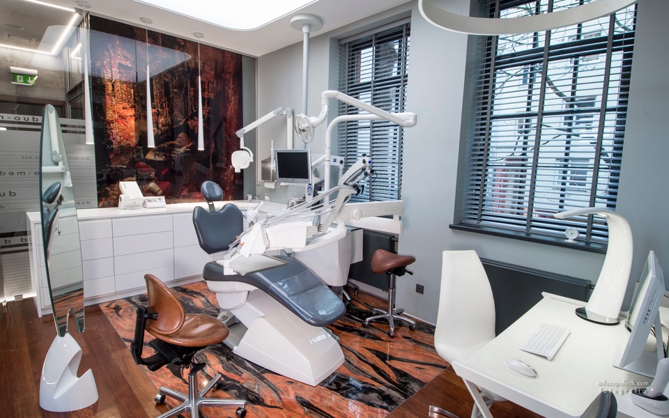 Nowoczesny gabinet stomatologiczny z fotelem dentystycznym, brązowym krzesłem na kółkach, białym biurkiem, komputerem i oknem z żaluzjami, na podłodze wzorzyste brązowo-czarne płytki.