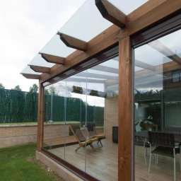 PARTNERHOUSE - Zadaszony taras z drewnianą konstrukcją, przeszklonymi ścianami przesuwnymi i widocznym ogrodem w tle.