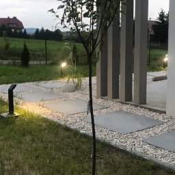 PARTNERHOUSE - Nowoczesna ścieżka z betonowych płyt i białego żwiru prowadząca do domu, oświetlona lampami ogrodowymi, w tle widok na dom i zieleń.