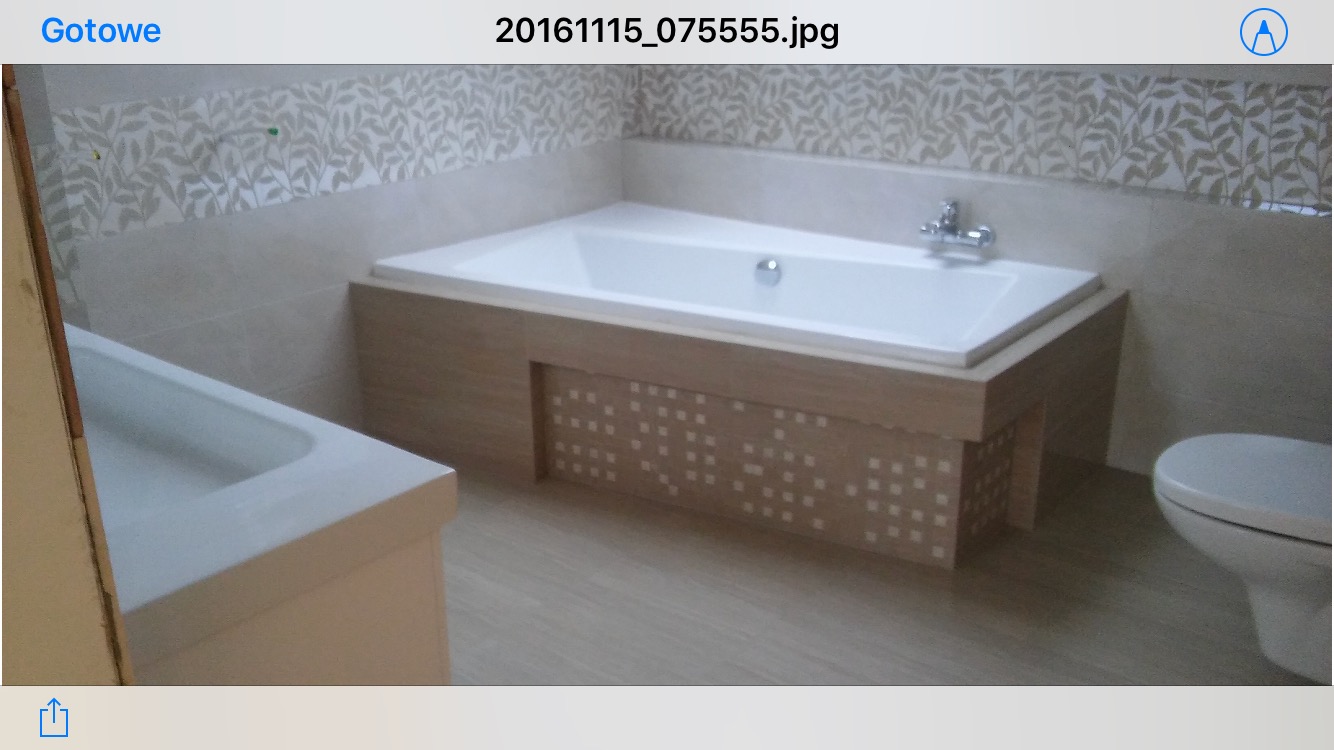 Wykończona łazienka z wanną obudowaną drewnopodobną płytą z ażurowym wzorem, widoczna również umywalka i toaleta, na ścianach płytki z subtelnym motywem roślinnym.