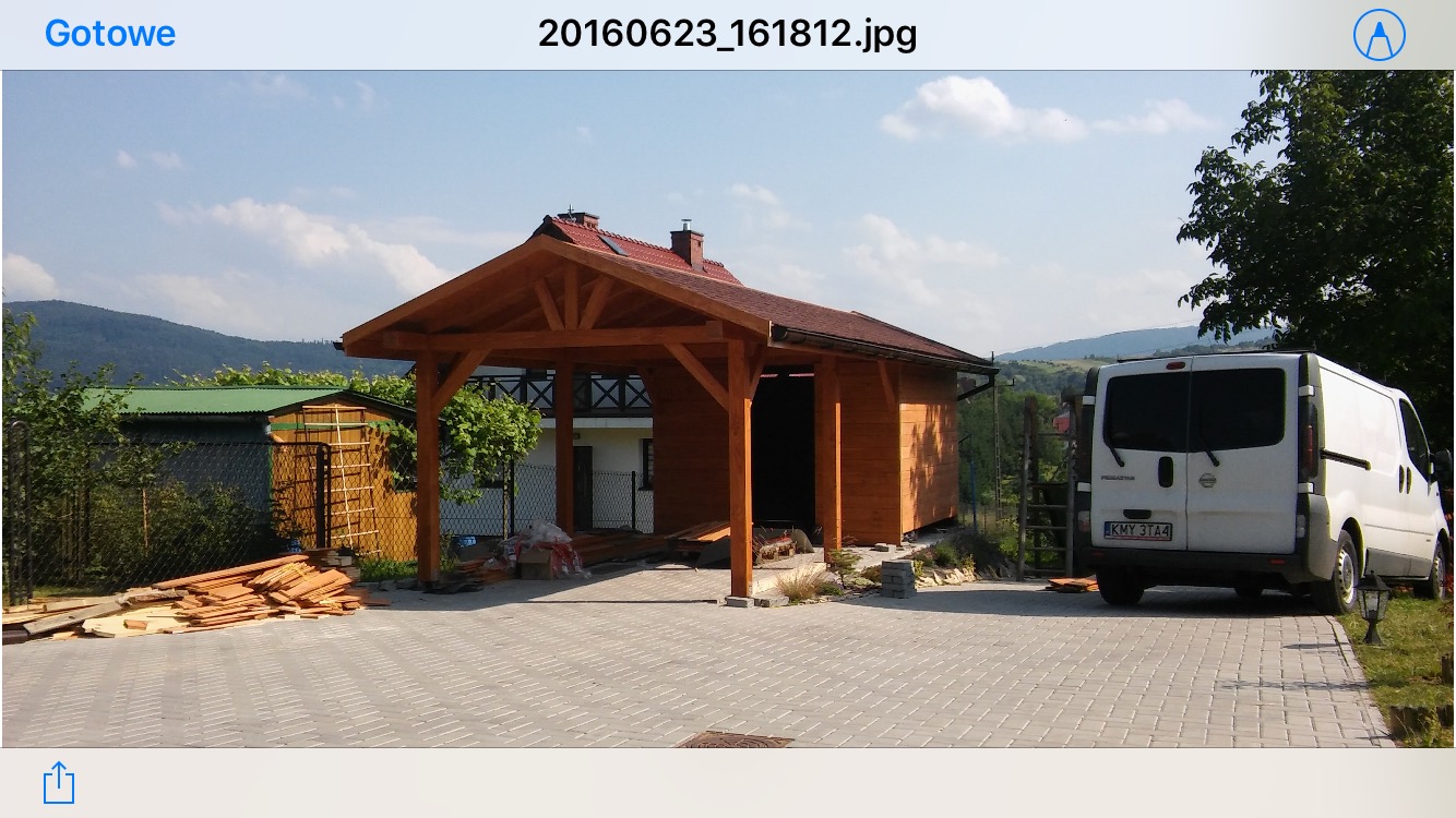 Drewniana altana z czerwonym dachem w trakcie budowy, widoczny stos desek na brukowanym podjeździe, biały bus w tle oraz zalesione wzgórza w oddali.