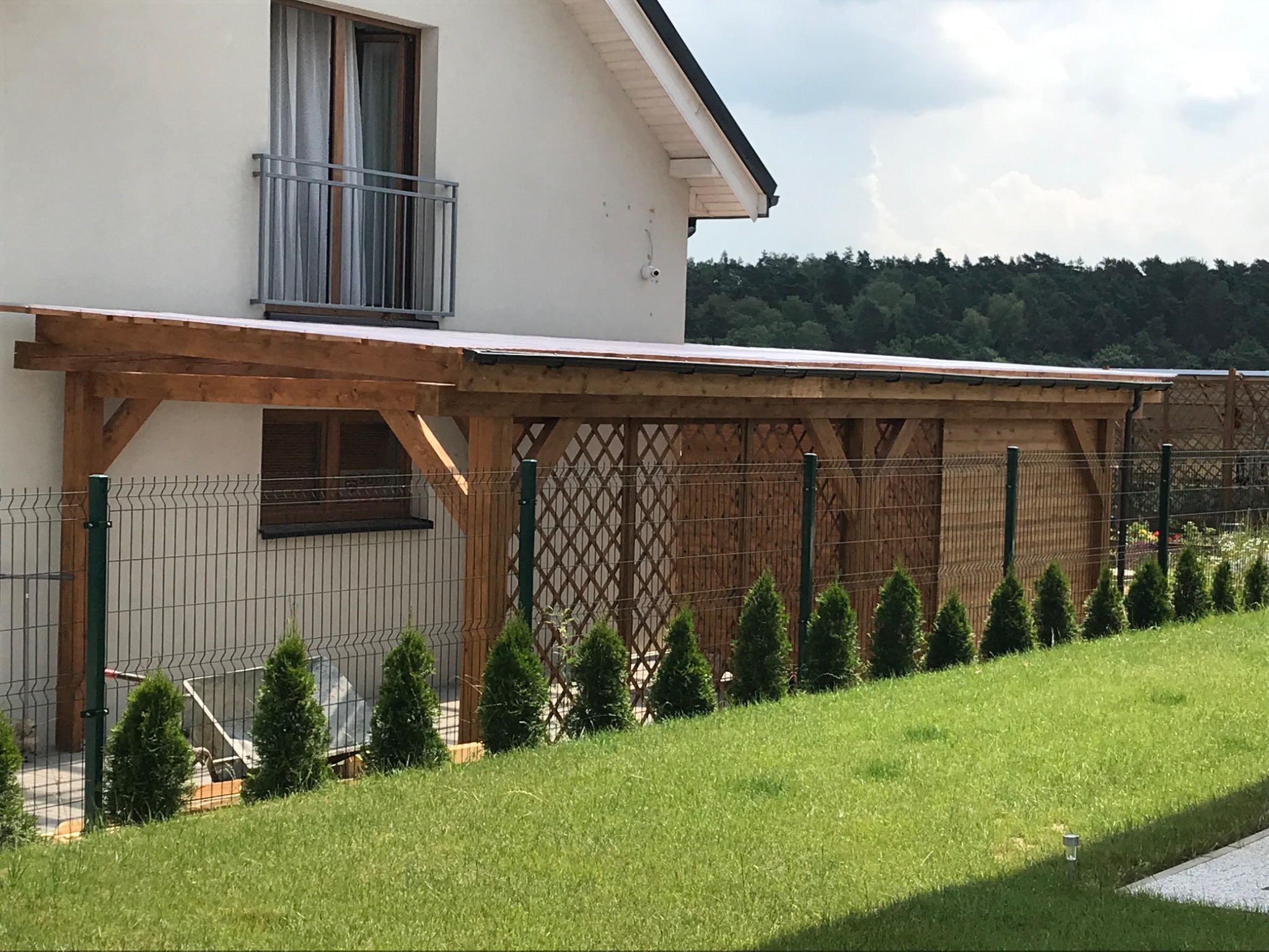Drewniana pergola z kratownicą przy białym domu jednorodzinnym, widok z trawnika, rząd małych tuj.