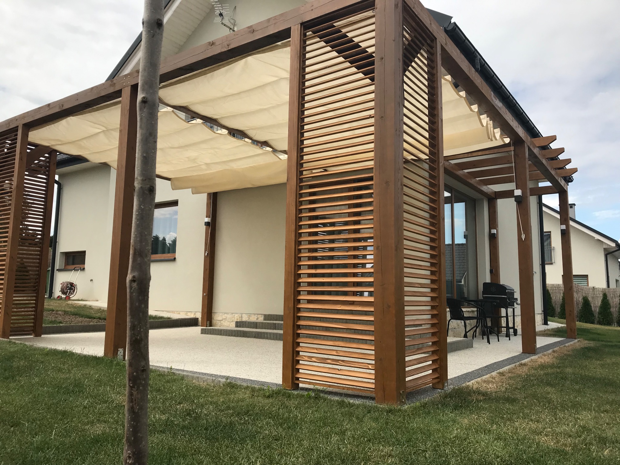 Drewniana pergola z beżową, zwijaną tkaniną przeciwsłoneczną, z poziomymi żaluzjami, na tarasie z szarego kamienia, z grillem i meblami ogrodowymi w tle.