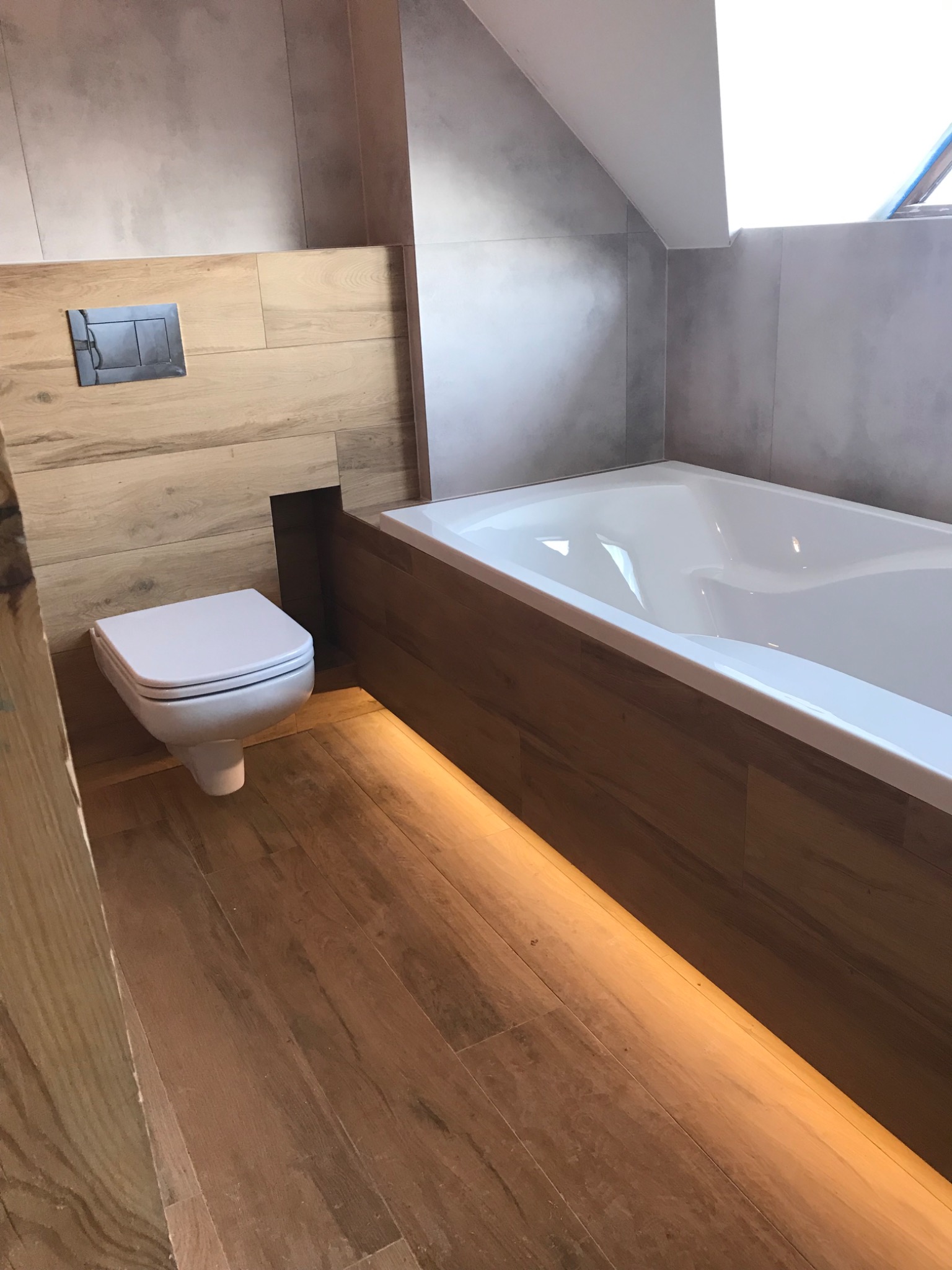 Nowoczesna łazienka z drewnopodobnymi płytkami, podświetleniem LED pod wanną i wiszącą toaletą z chromowanym przyciskiem spłukiwania.