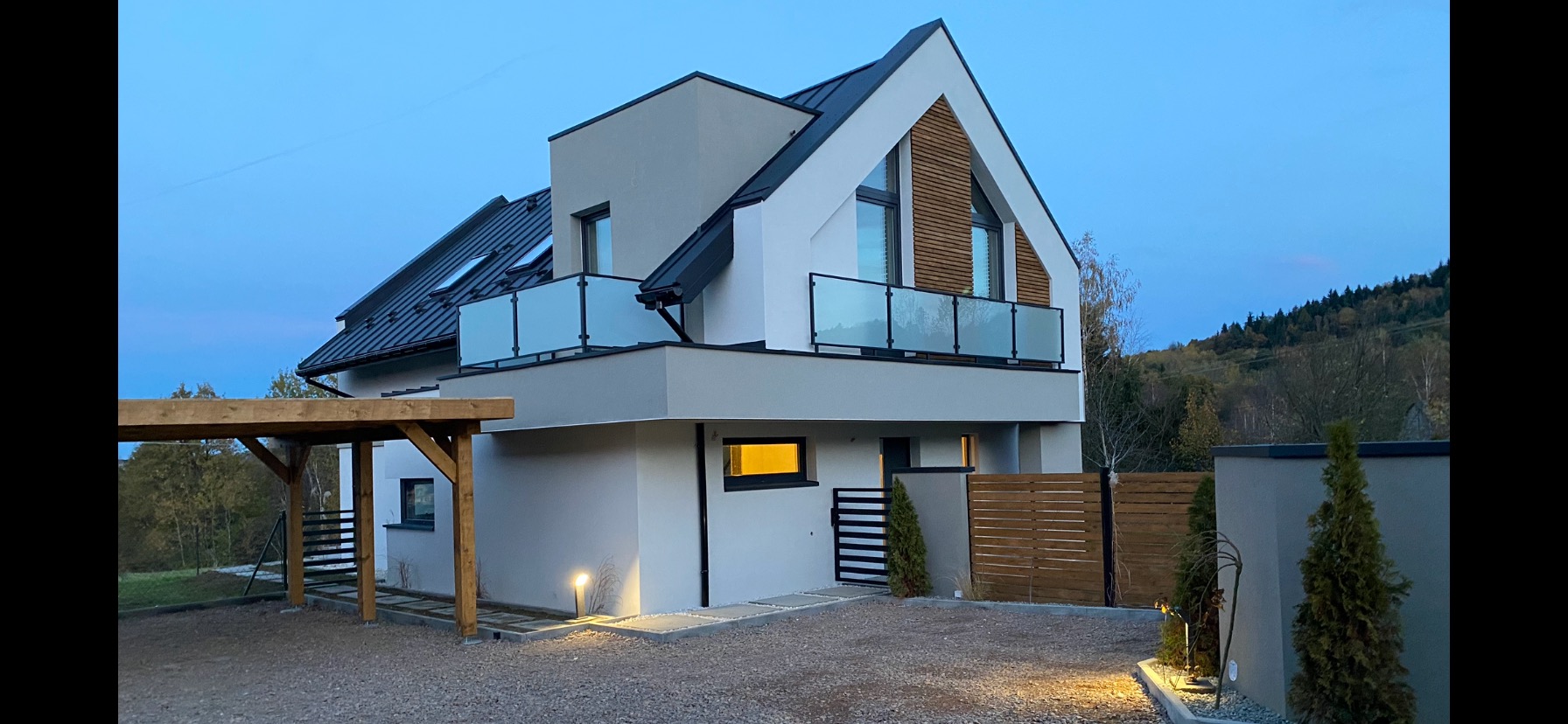 Nowoczesny dom jednorodzinny z drewnianą wiatą garażową i balkonem z szklaną balustradą, oświetlony lampami zewnętrznymi, na tle zalesionego wzgórza o zmierzchu.
