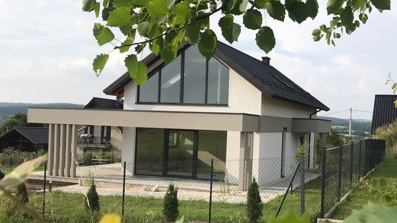 Nowoczesny dom jednorodzinny z dużymi przeszkleniami, wykończony w bieli i szarości, z czarnym dachem i minimalistycznym ogrodem, widok na pagórkowaty krajobraz w tle.