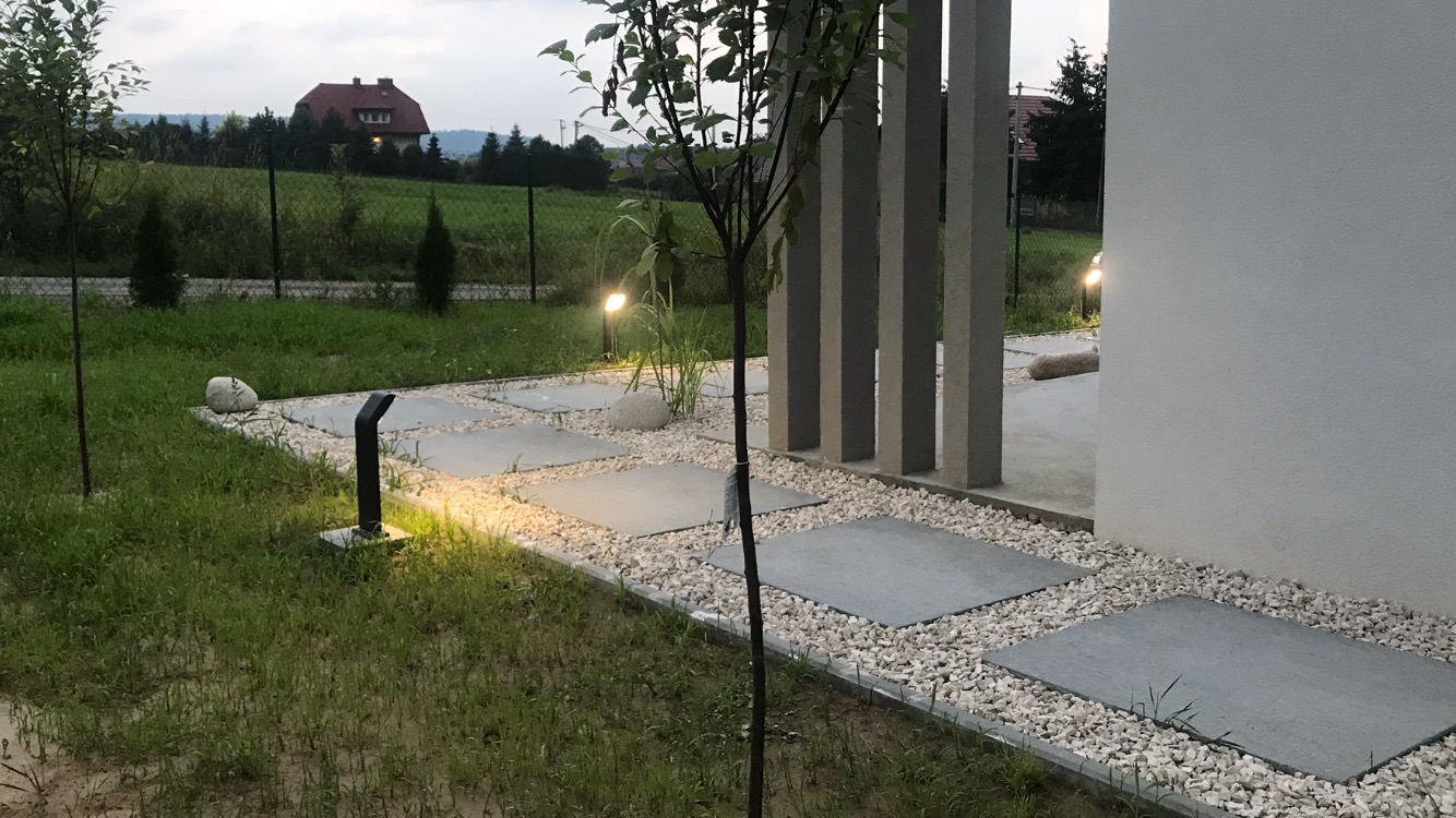 Nowoczesna ścieżka z betonowych płyt i białego żwiru prowadząca do domu, oświetlona lampami ogrodowymi, w tle widok na dom i zieleń.