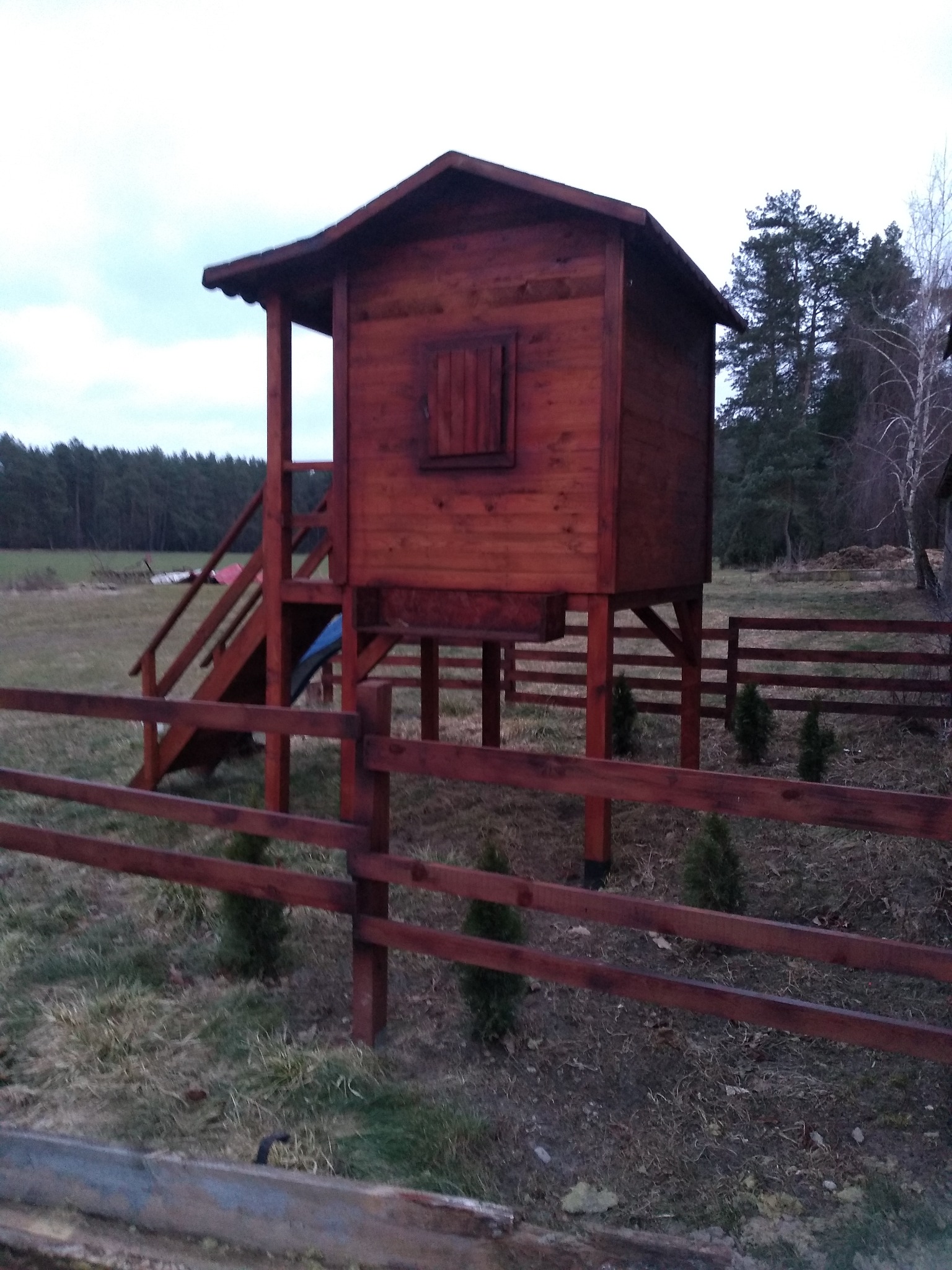 Drewniany domek na podwyższeniu z drabinką i zjeżdżalnią, otoczony niskim płotem, widoczny na tle łąki i lasu.