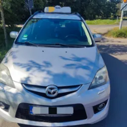 Srebrna Mazda z napisem TAXI na dachu, zaparkowana przy drodze, widok z przodu, tablica rejestracyjna z Polski.