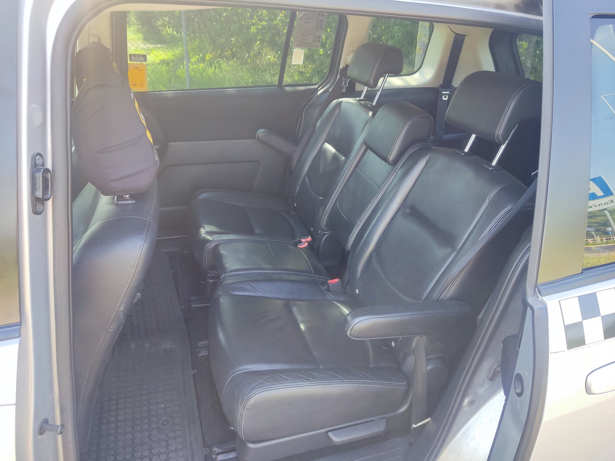 Wnętrze samochodu typu van z trzema rzędami skórzanych foteli, widoczna naklejka z cennikiem i logo 'Blik' na szybie, oraz szachownica na karoserii z tyłu.