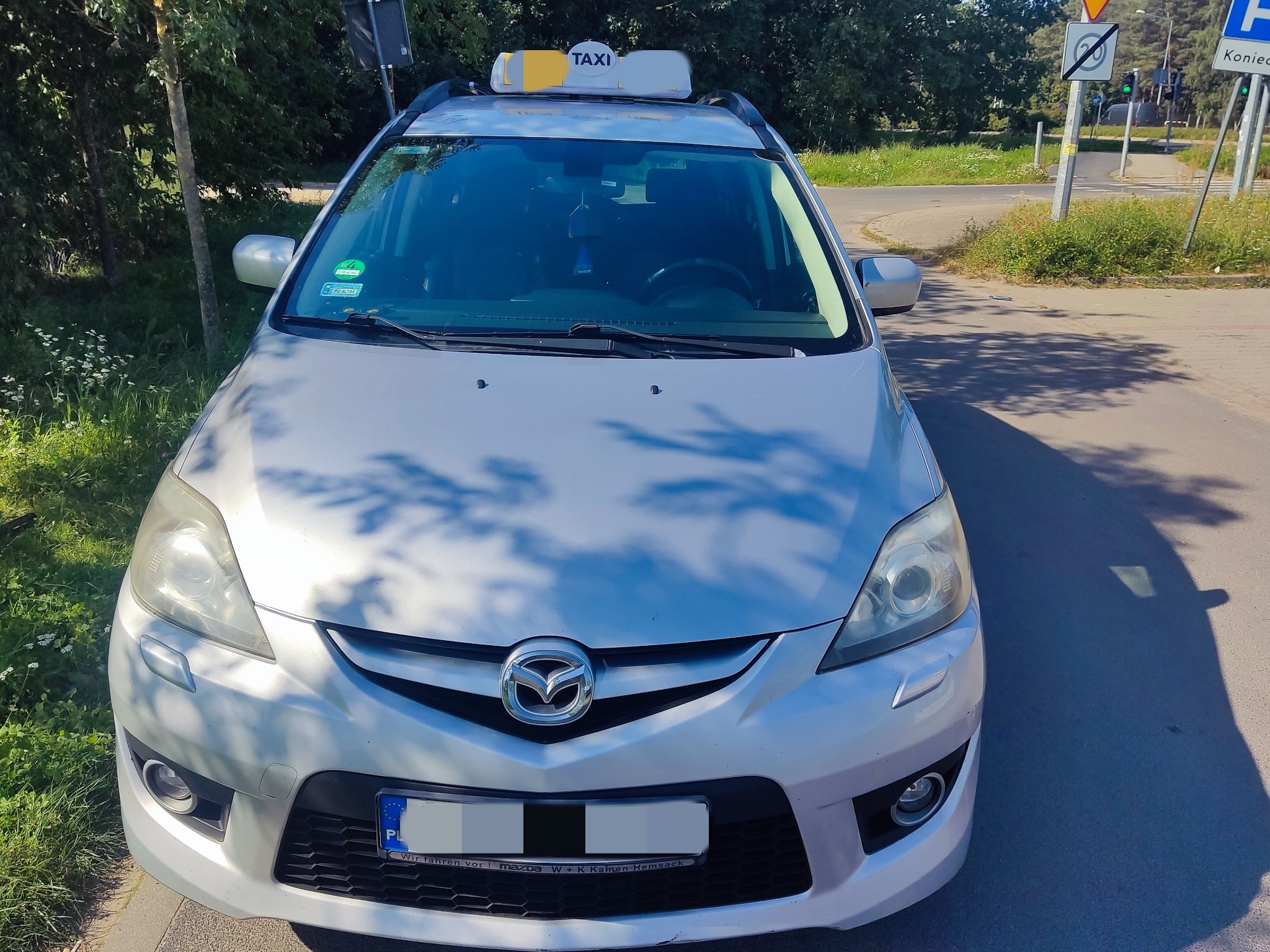 Srebrna Mazda z napisem TAXI na dachu, zaparkowana przy drodze, widok z przodu, tablica rejestracyjna z Polski.