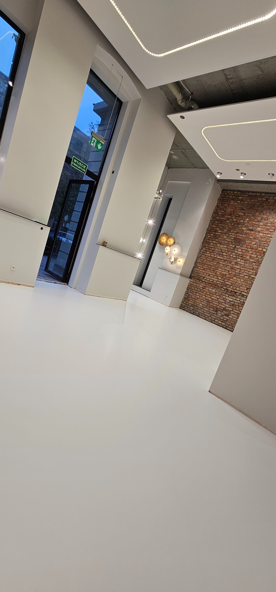 Jasne, przestronne wnętrze z białą posadzką żywiczną, ceglaną ścianą akcentującą, nowoczesnym oświetleniem sufitowym LED i minimalistycznymi dekoracjami ściennymi.