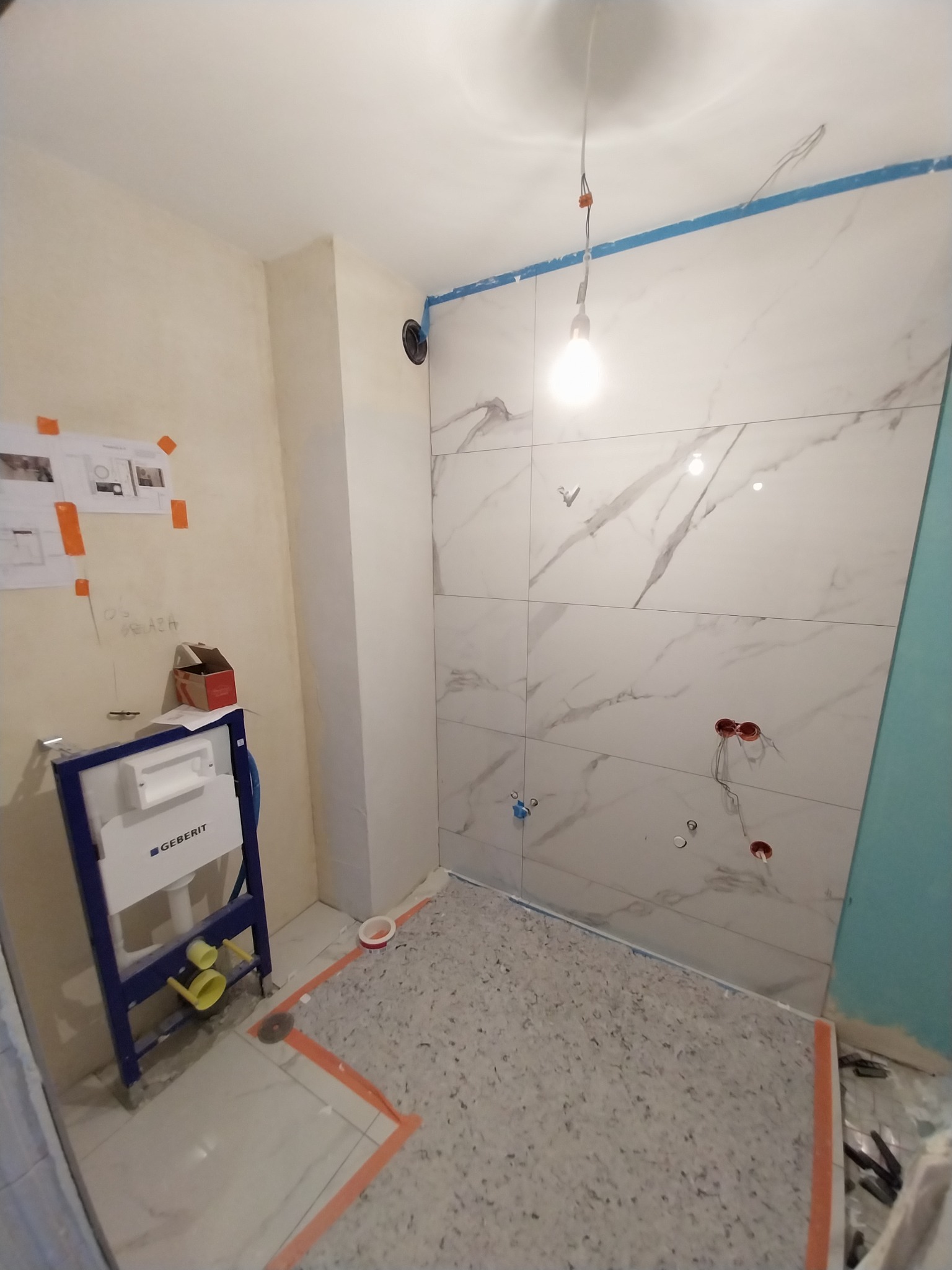 Łazienka w trakcie prac: białe płytki imitujące marmur na ścianach, widoczna instalacja podtynkowego WC Geberit, zabezpieczona podłoga, wystające przewody elektryczne i rury wodne.