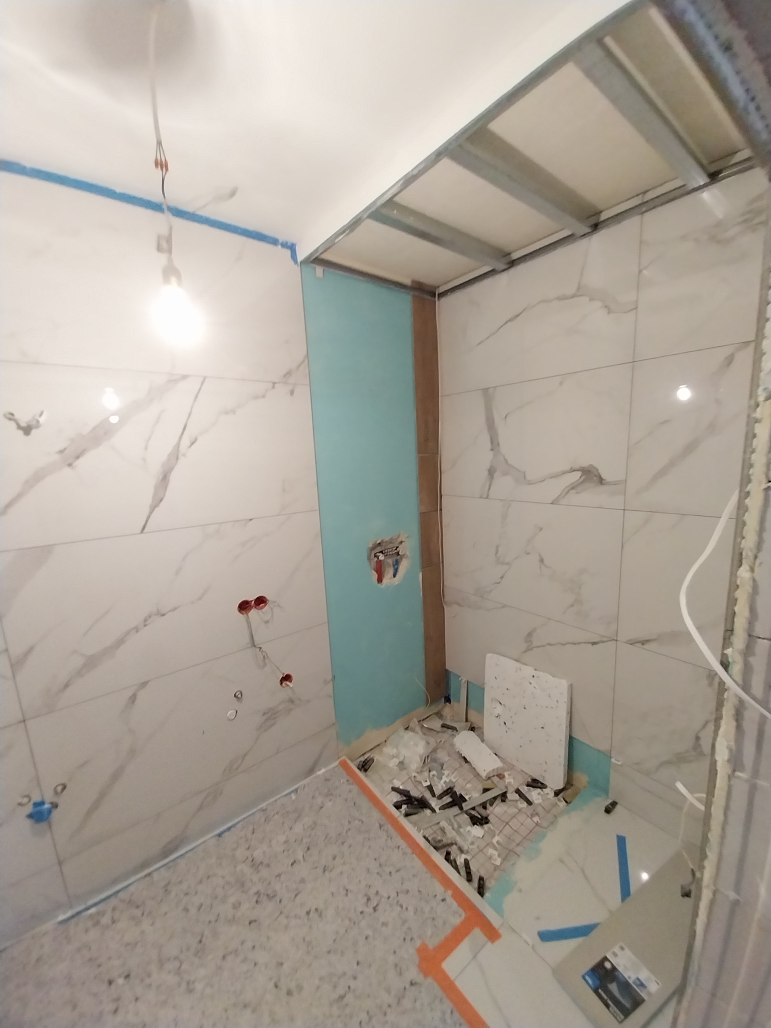 Wnętrze łazienki w trakcie remontu: położone płytki imitujące marmur na ścianach, widoczne profile sufitowe, fragment niebieskiej ściany, narzędzia i akcesoria glazurnicze na podłodze. Częściowo...