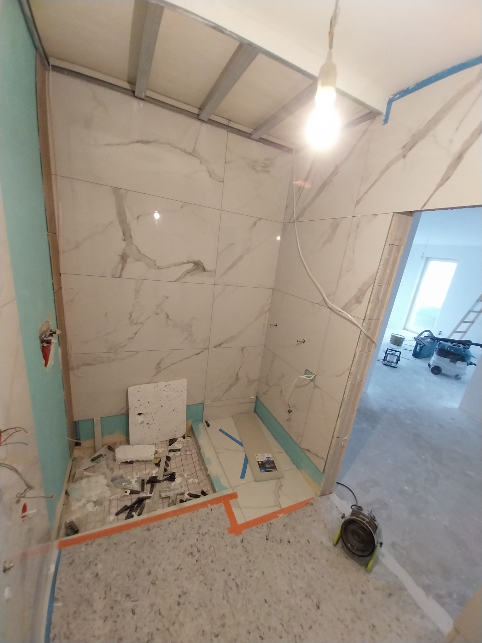 Łazienka w trakcie układania płytek marmuropodobnych na ścianach, widoczny system ogrzewania podłogowego, fragment płyty gipsowej, surowa żarówka oświetlająca wnętrze i narzędzia pracy.