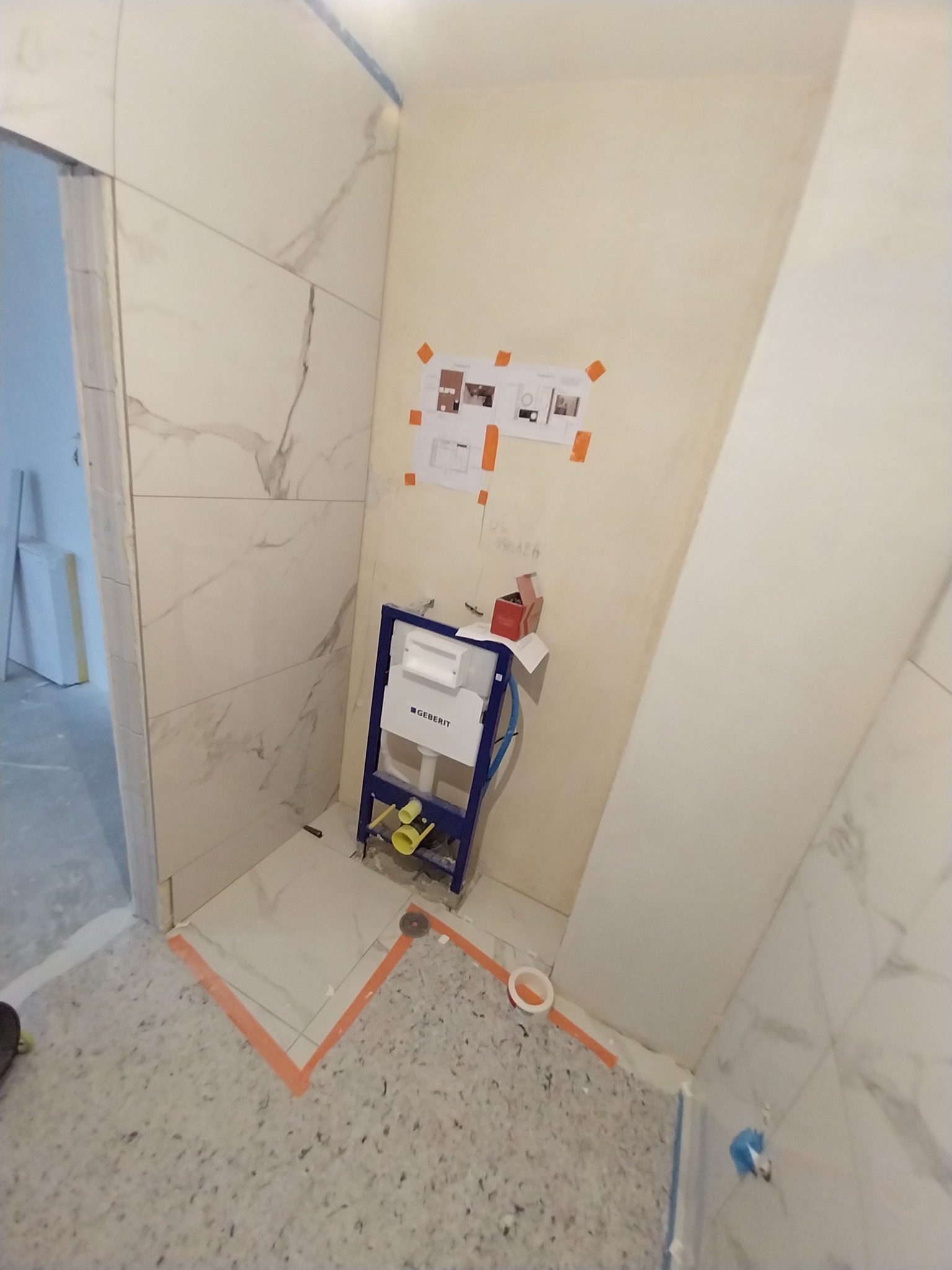Łazienka w trakcie remontu: białe płytki imitujące marmur na ścianach, stelaż podtynkowy Geberit zamontowany, widoczne plany i szkice na ścianie, przygotowanie do montażu ceramiki sanitarnej.