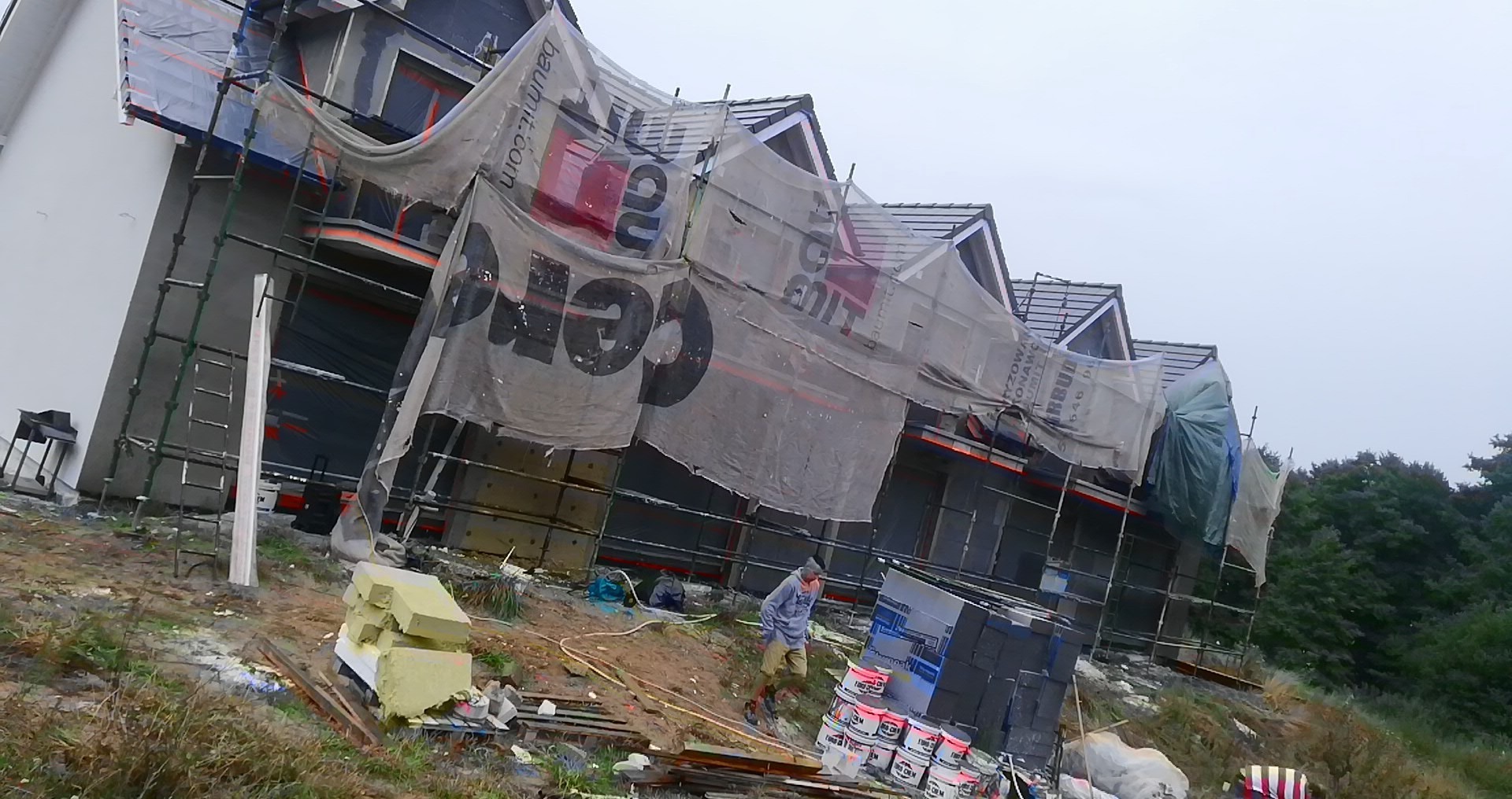 Dom w trakcie prac elewacyjnych, osłonięty siatką z nadrukiem reklamowym, widoczne rusztowania, materiały budowlane i pracownik na terenie budowy.