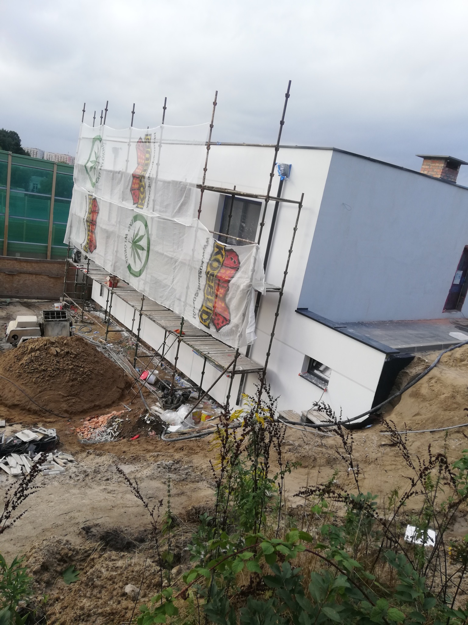 Nowoczesny dom w trakcie prac elewacyjnych, rusztowanie z siatką osłonową z nadrukiem, widoczny stos materiałów budowlanych i teren wokół w trakcie przygotowania.