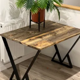 Stół z blatem z postarzanego drewna i czarnymi, metalowymi nogami w kształcie litery X, obok biały grzejnik, na stole doniczka z zieloną rośliną.