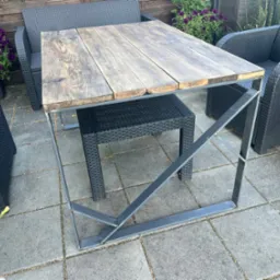 Stół ogrodowy z blatem z desek i stalową, spawaną konstrukcją nóg, otoczony rattanowymi krzesłami i kwiatami w donicach na tarasie.