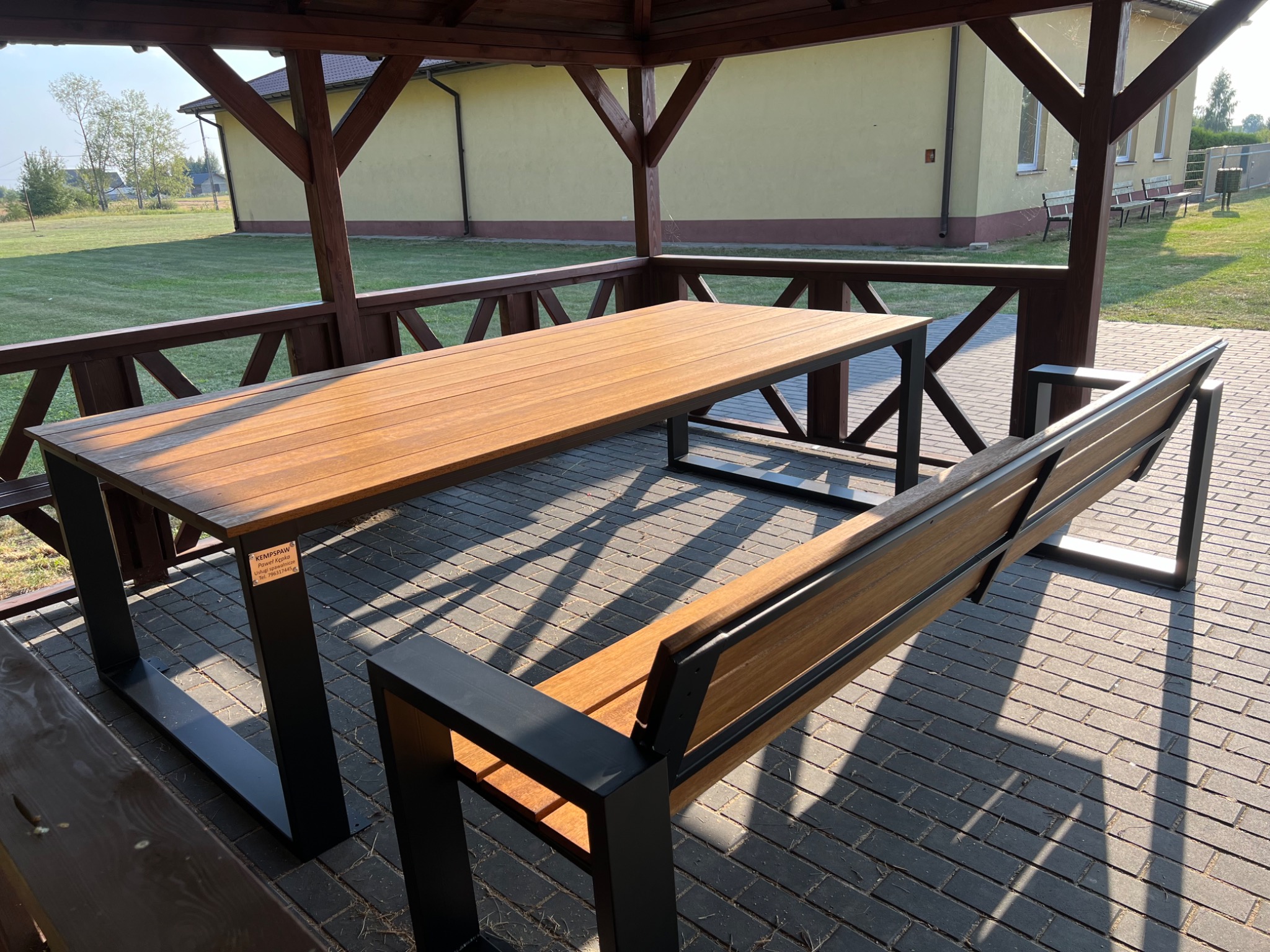 Stół i ławka z drewnianym blatem i siedziskiem oraz metalową konstrukcją w altanie, widoczne cienie i budynek w tle.