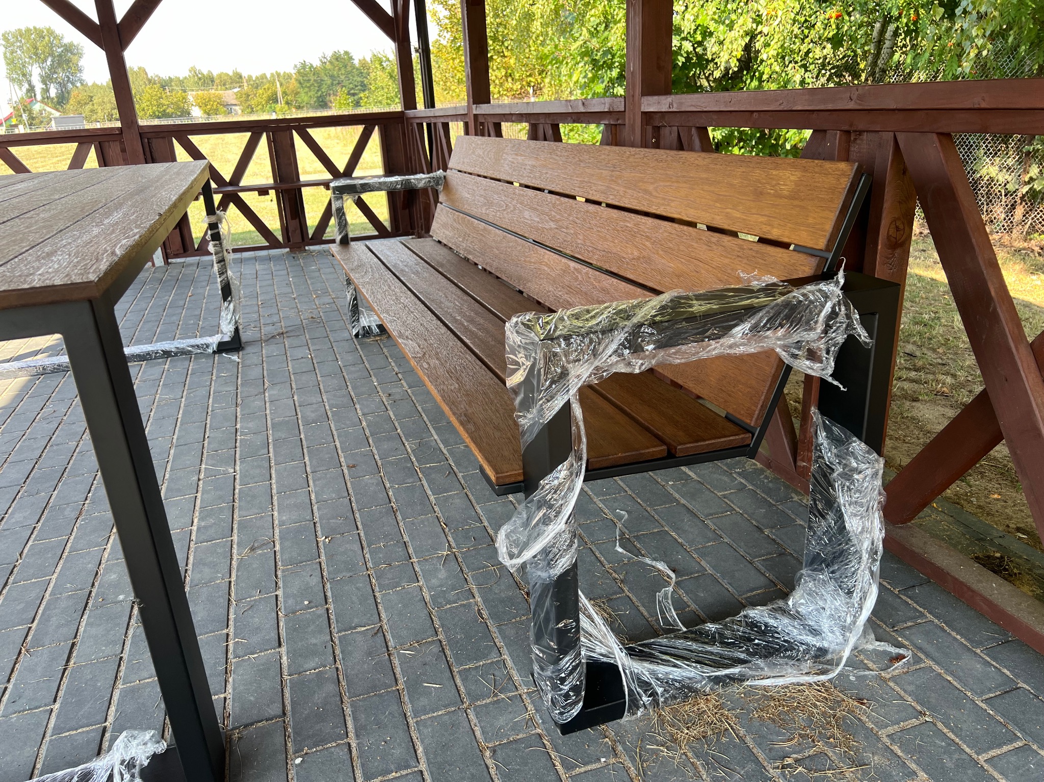 Nowy drewniany stół i ławka z czarnymi metalowymi nogami, jeszcze w folii ochronnej, ustawione w drewnianej altanie z widokiem na zieleń.