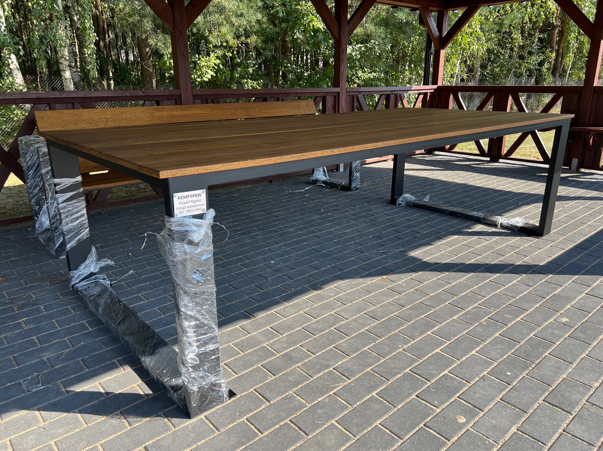 Stół ogrodowy z drewnianym blatem i metalową ramą, częściowo owinięty folią ochronną, stojący w altanie na kostce brukowej.