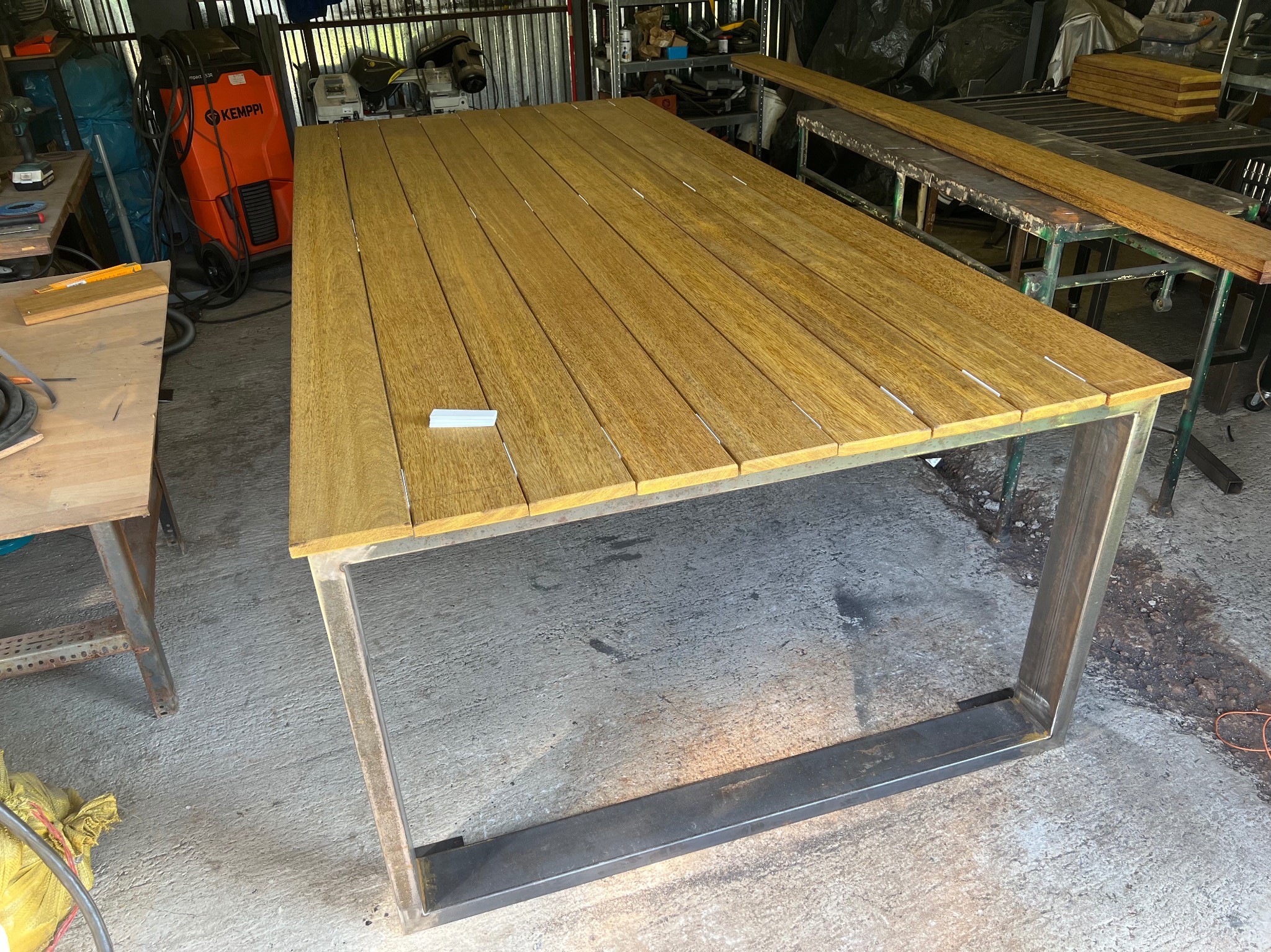 Stół z drewnianym blatem o ciepłym odcieniu i metalową ramą w warsztacie, widoczny sprzęt spawalniczy w tle.