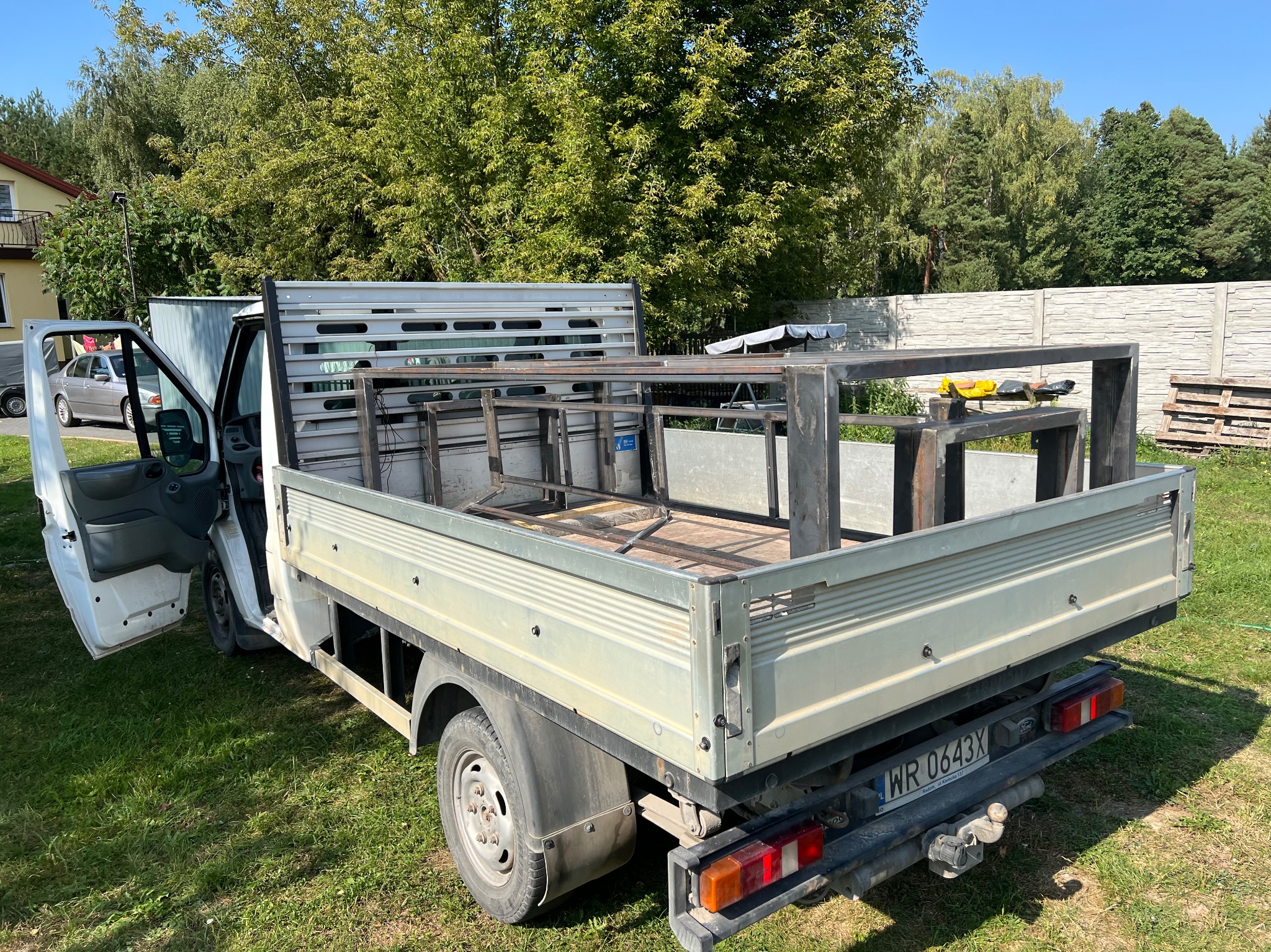 Biały samochód dostawczy z otwartymi drzwiami i konstrukcją ze spawanych metalowych profili w skrzyni ładunkowej, widok z tyłu na tle zieleni.