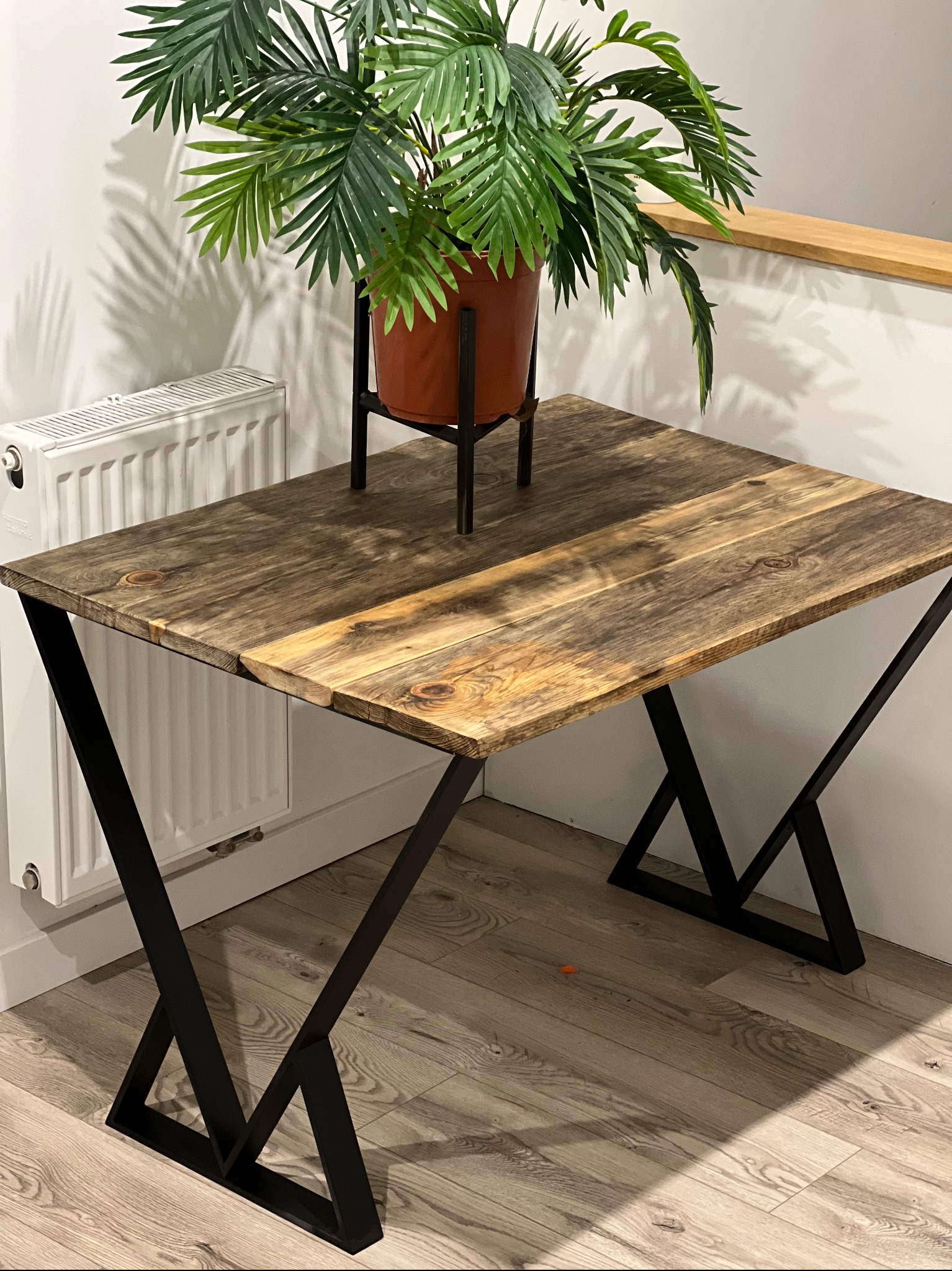 Stół z blatem z postarzanego drewna i czarnymi, metalowymi nogami w kształcie litery X, obok biały grzejnik, na stole doniczka z zieloną rośliną.