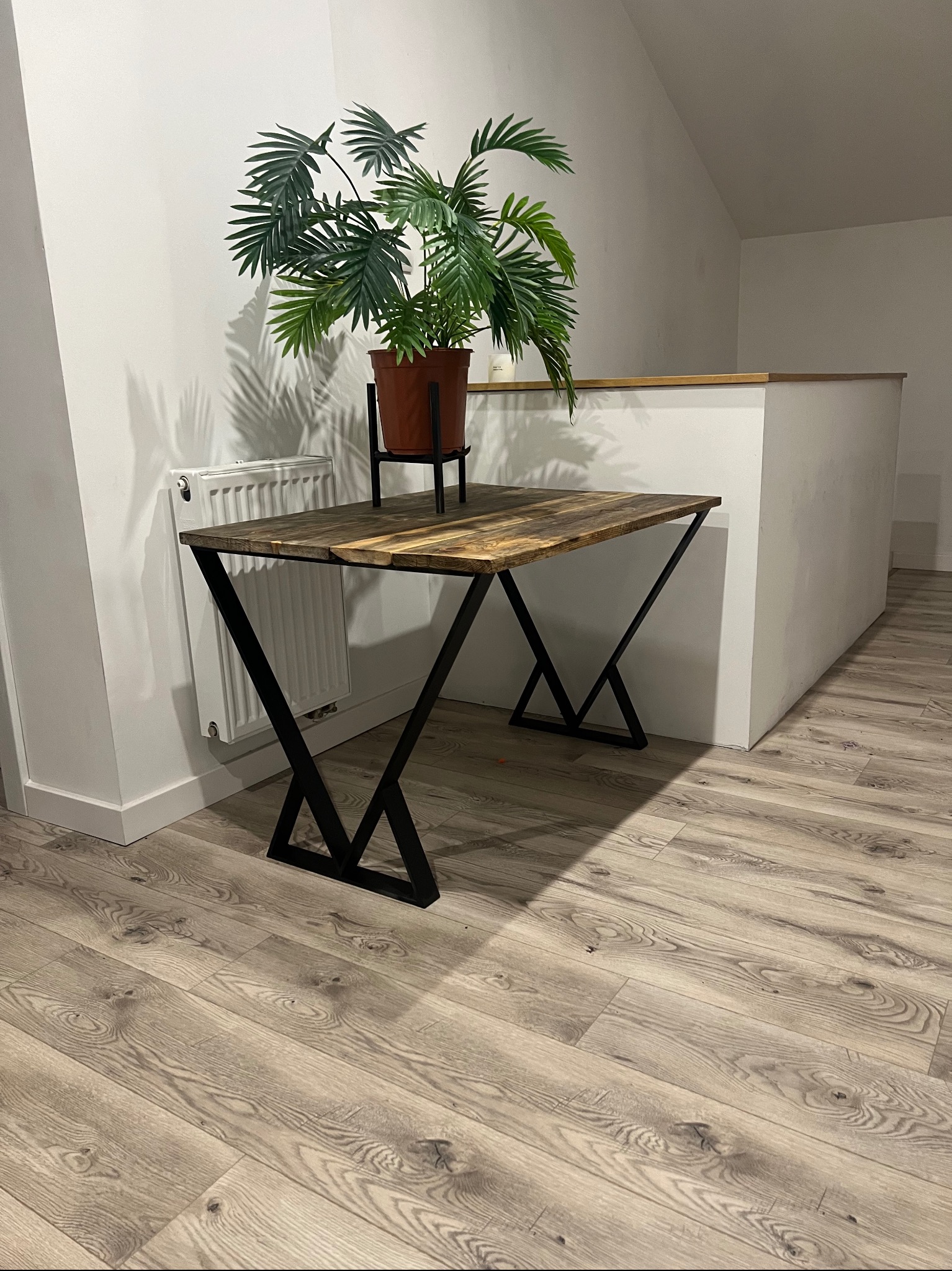 Stół z blatem z ciemnego drewna i czarnymi, metalowymi nogami w kształcie litery X, na stole doniczka z zieloną rośliną, w tle biała ściana i grzejnik.