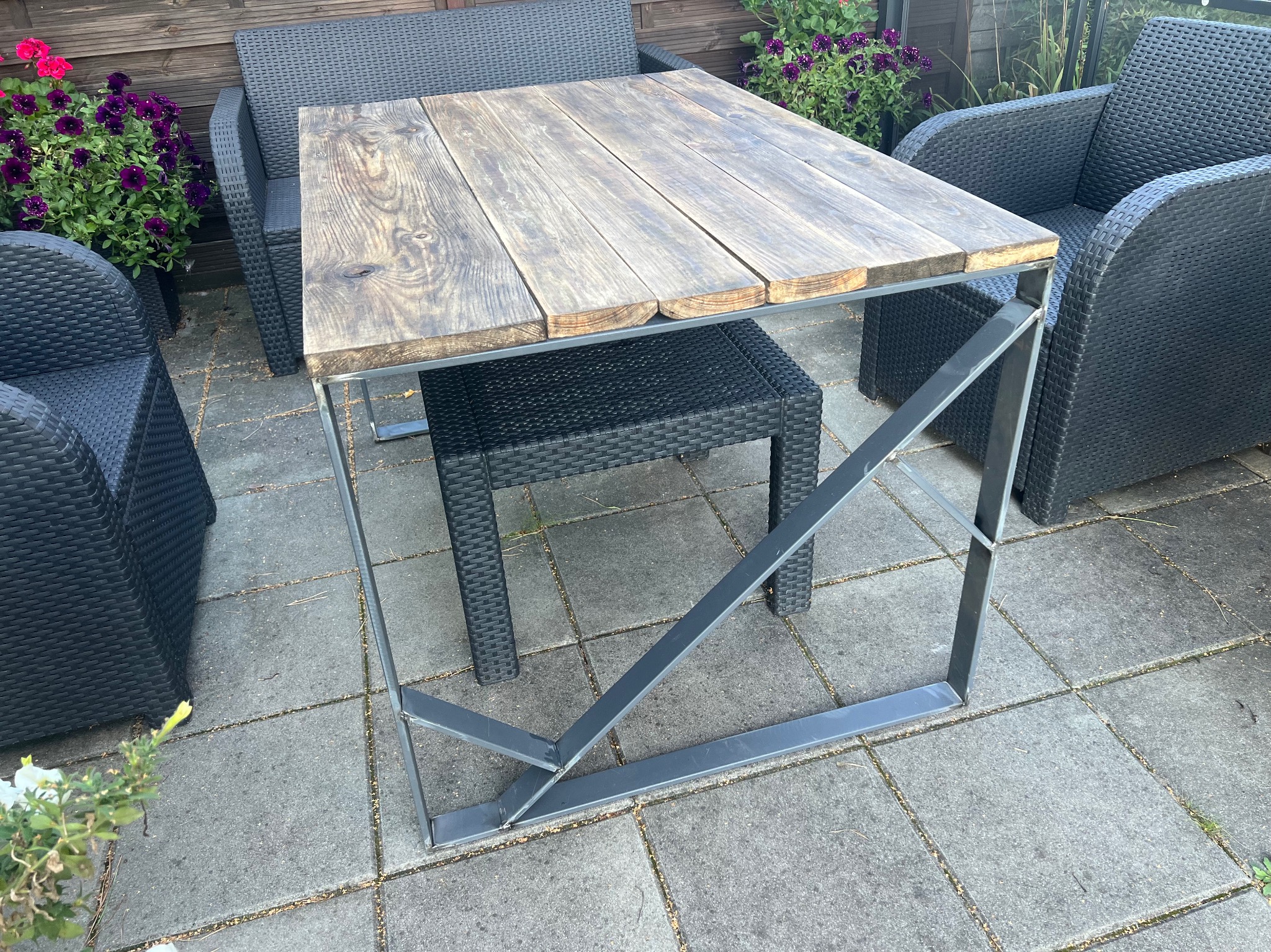 Stół ogrodowy z blatem z desek i stalową, spawaną konstrukcją nóg, otoczony rattanowymi krzesłami i kwiatami w donicach na tarasie.