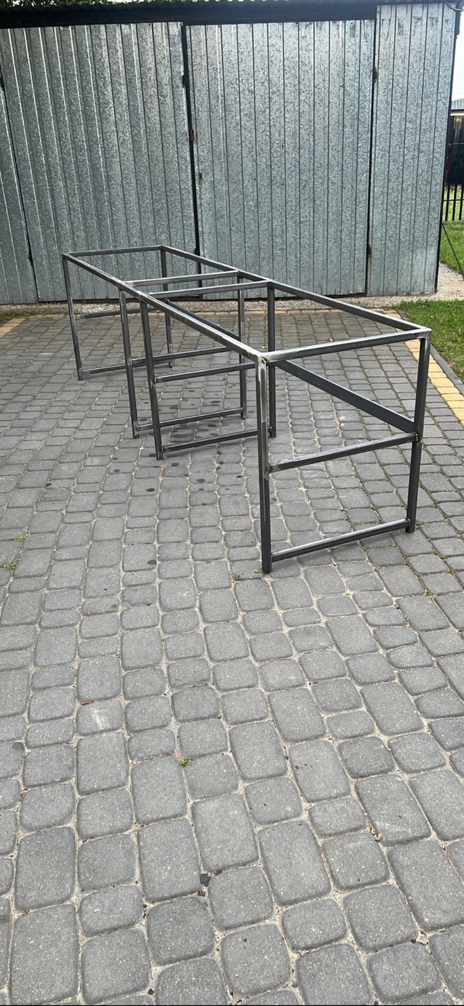 Surowa, spawana konstrukcja stalowa z profili, prawdopodobnie rama stołu lub innego mebla, na tle metalowego garażu i kostki brukowej.