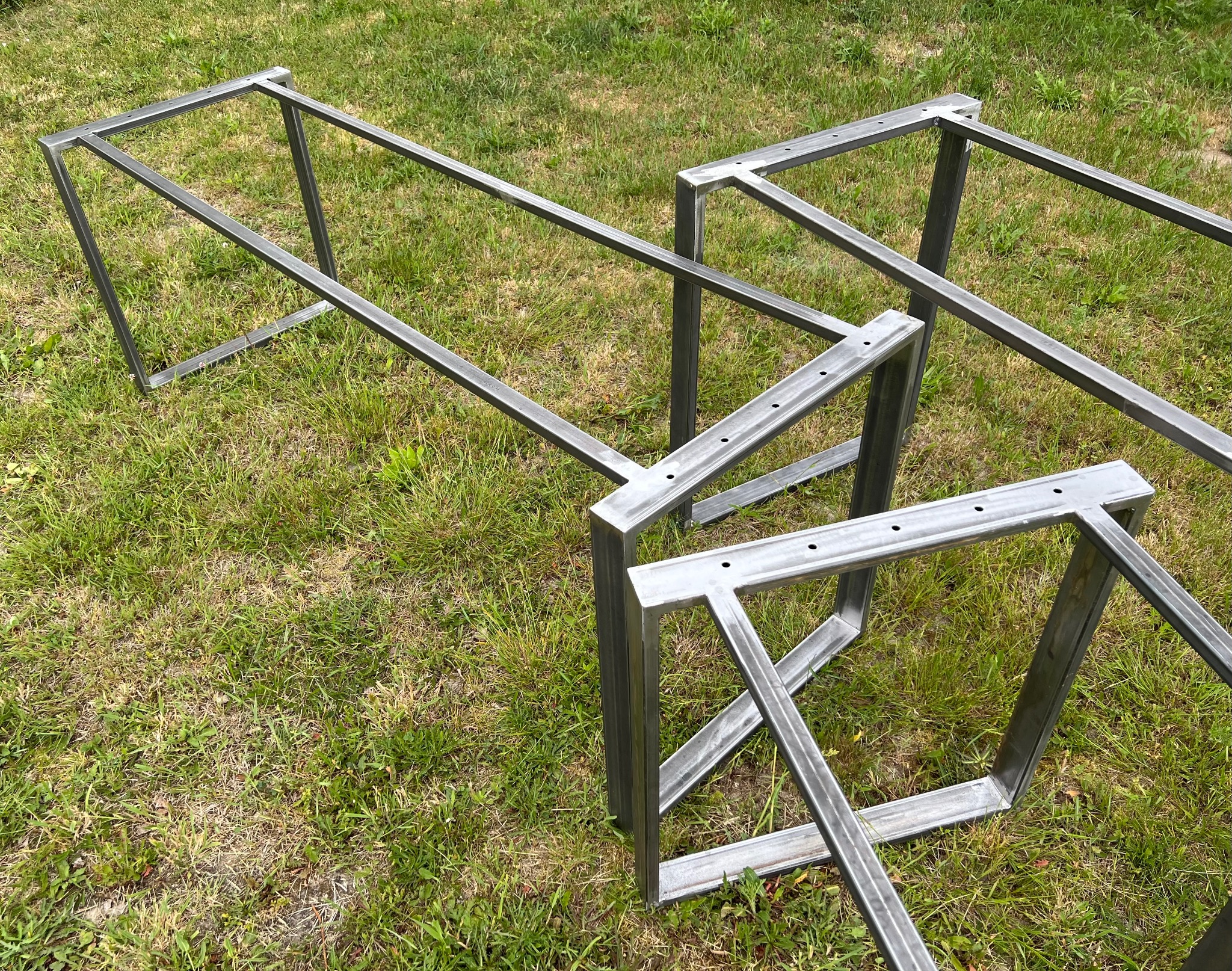 Surowe, spawane metalowe ramy stołów z otworami na śruby, leżące na trawie.