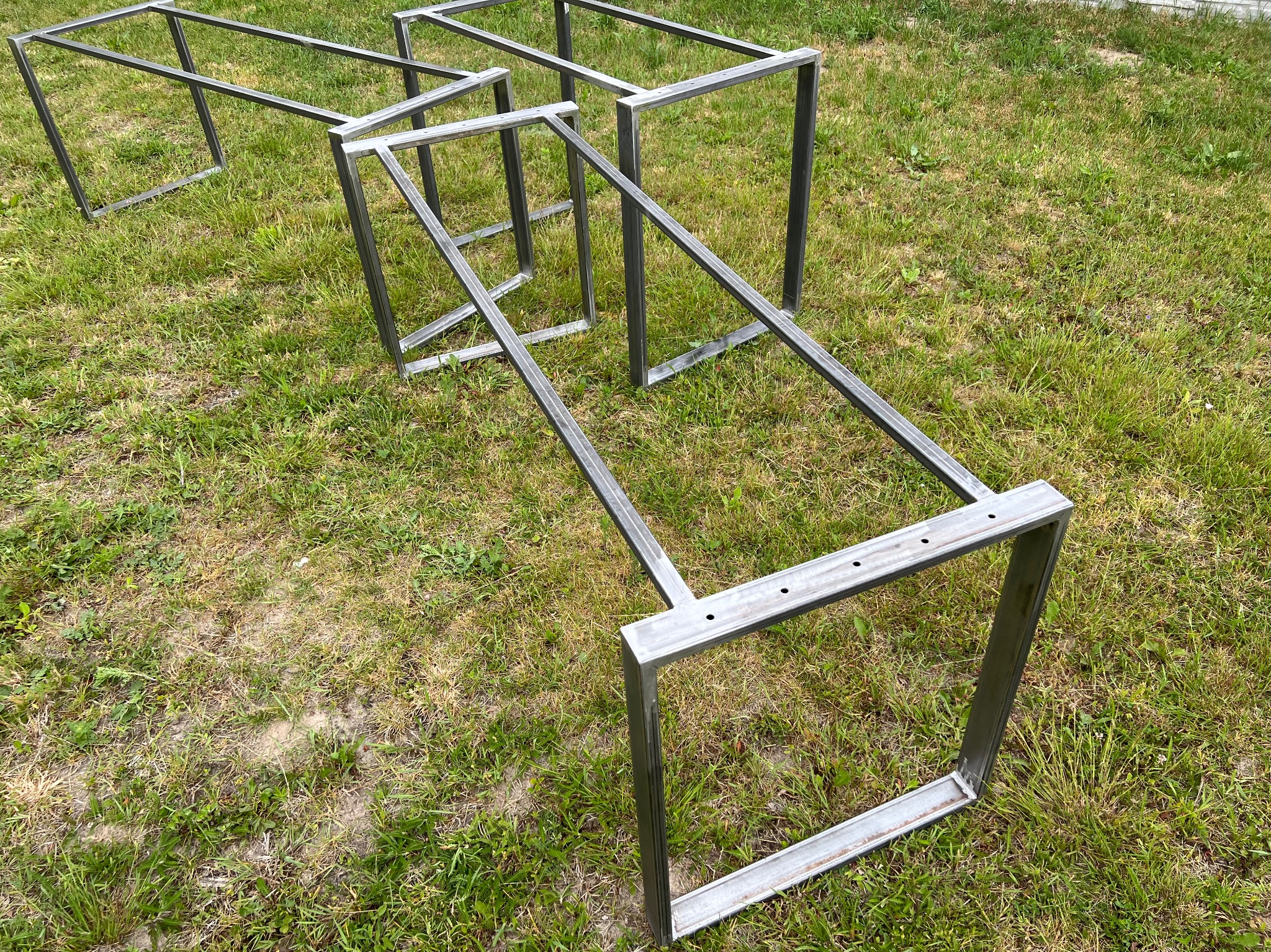 Surowe, metalowe ramy stołów lub podstawy mebli na trawie, widoczne spawy i otwory montażowe.